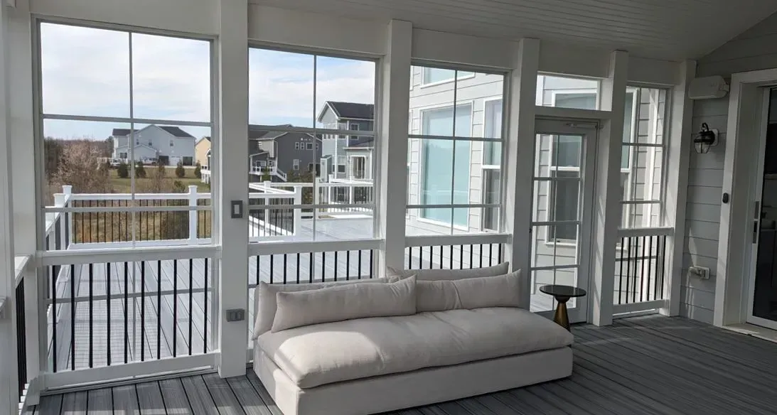 A white couch sits inside a screened porch with a view of a snowy backyard and other houses.