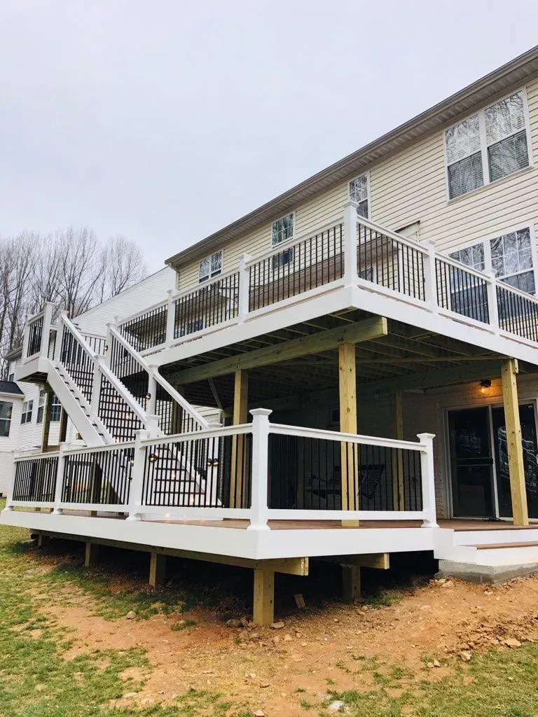 Two-story deck on a house, white railing, stairs to the ground. Beige siding, cloudy sky.