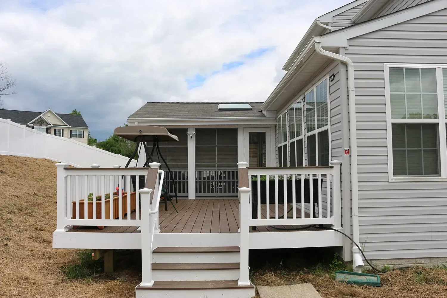 A deck with white railings and steps leads to a screened porch attached to a house with gray siding.