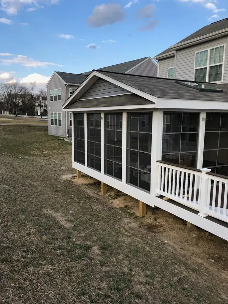 Screened porch with white railings and roof, attached to a gray house, on a grassy hill.