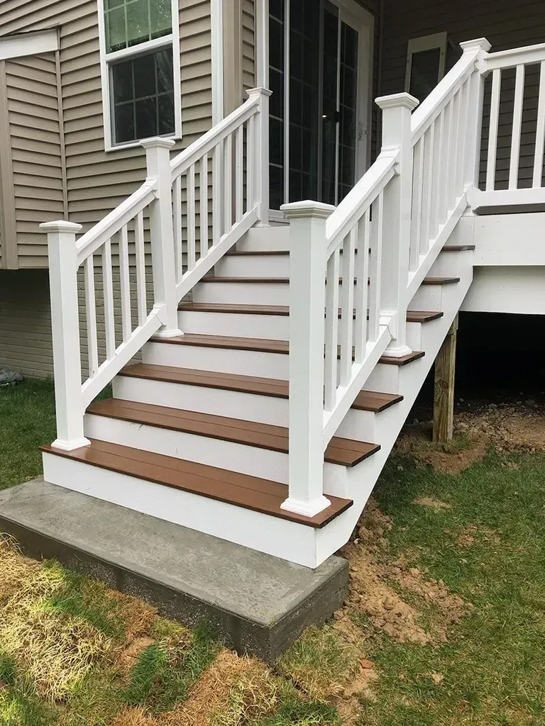 White-railed outdoor stairs with brown steps leading up to a deck. They are beside a light-colored house.
