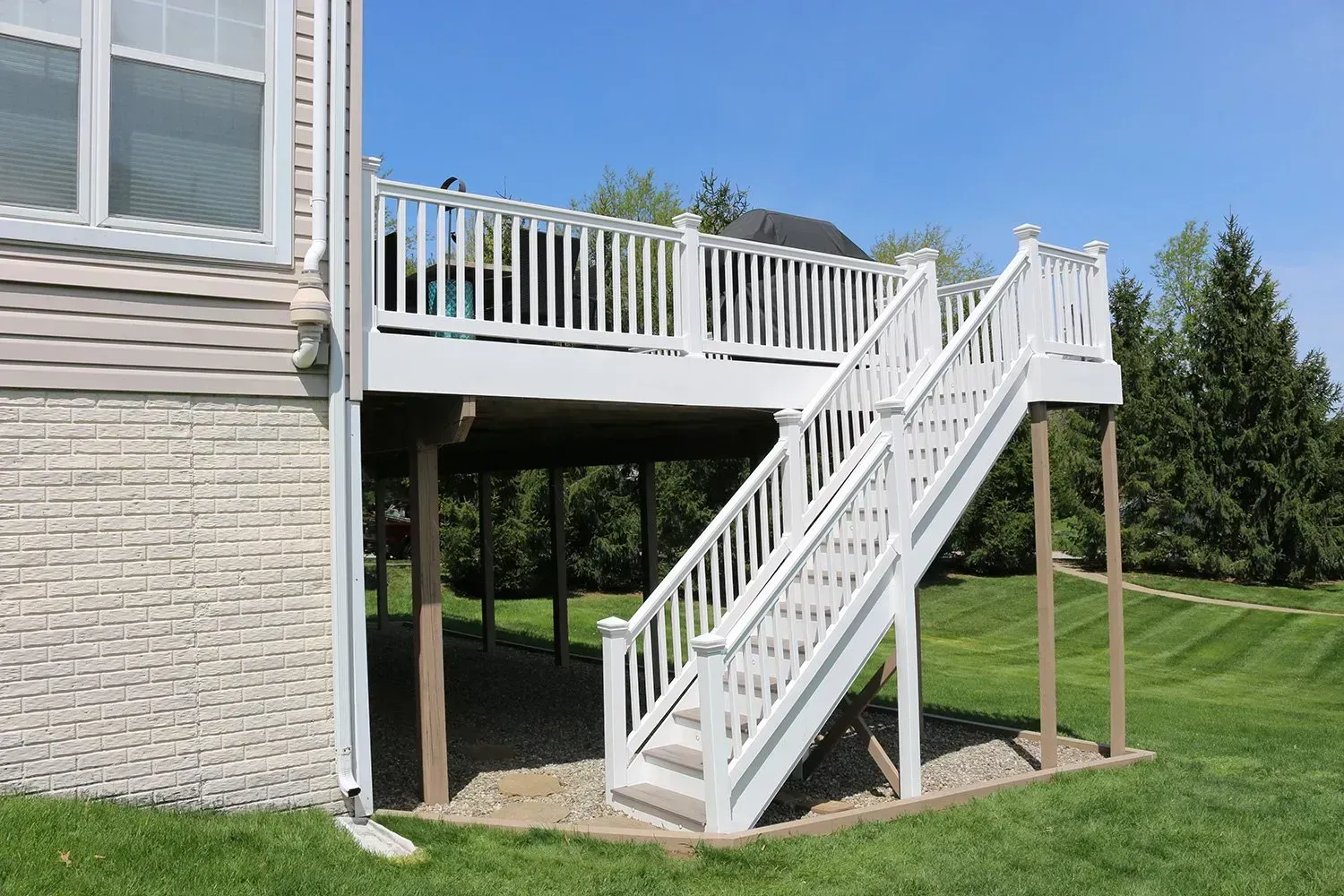 White elevated deck with stairs in a backyard. The deck is attached to a beige house, under a clear, blue sky.