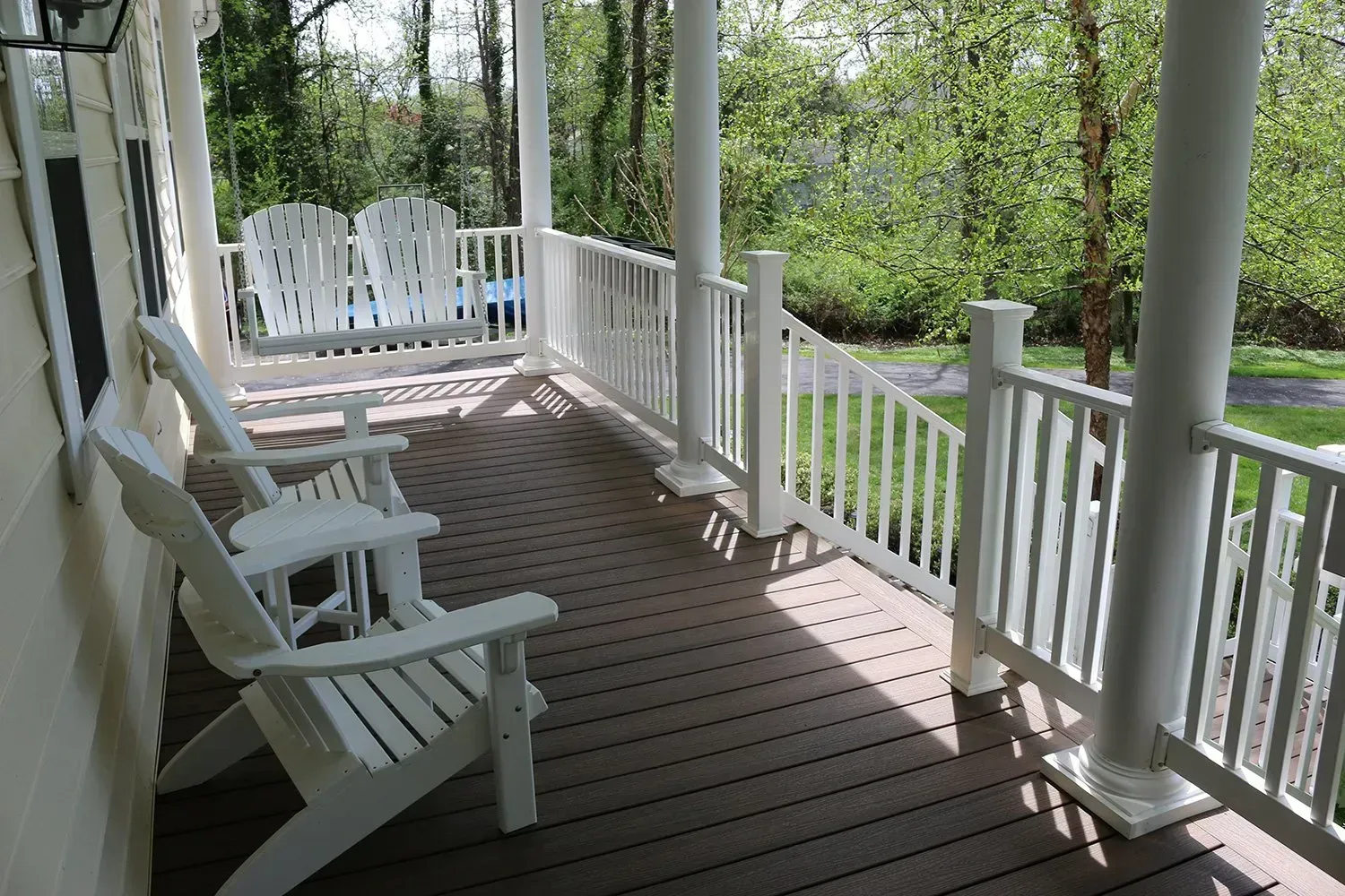 White chairs on a porch with white railing and columns, set against a backdrop of green trees.