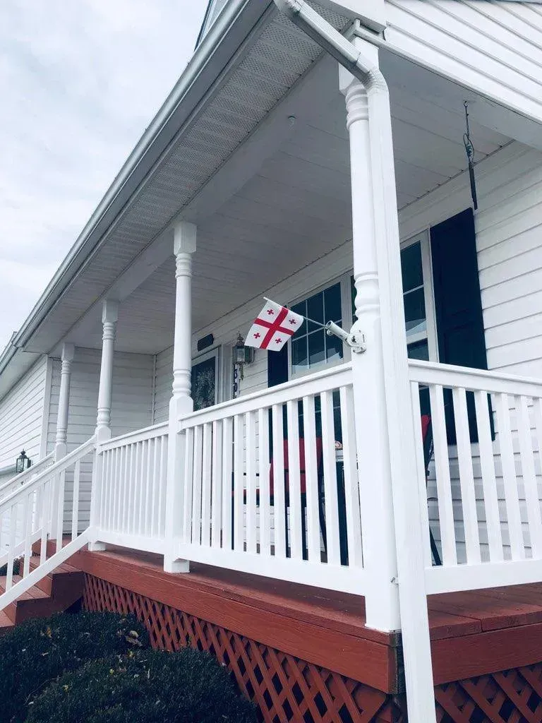 White house with porch, railing, red steps; a flag with a red cross on white background is displayed.