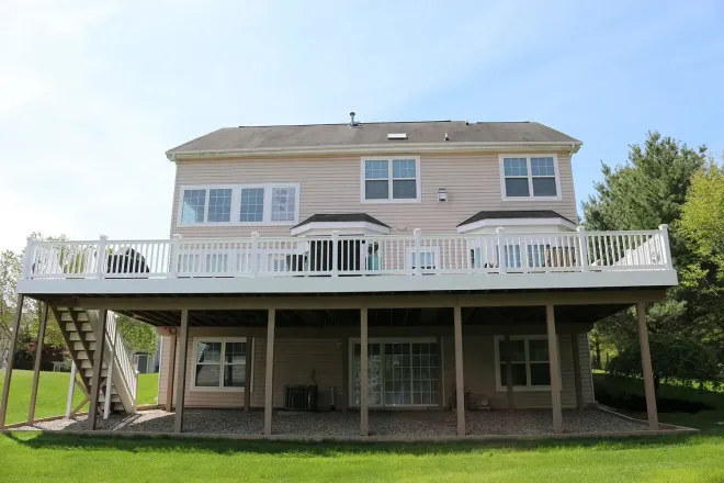 Two-story house with a large deck, white railing, and stairs. Beige siding, windows, and green lawn.