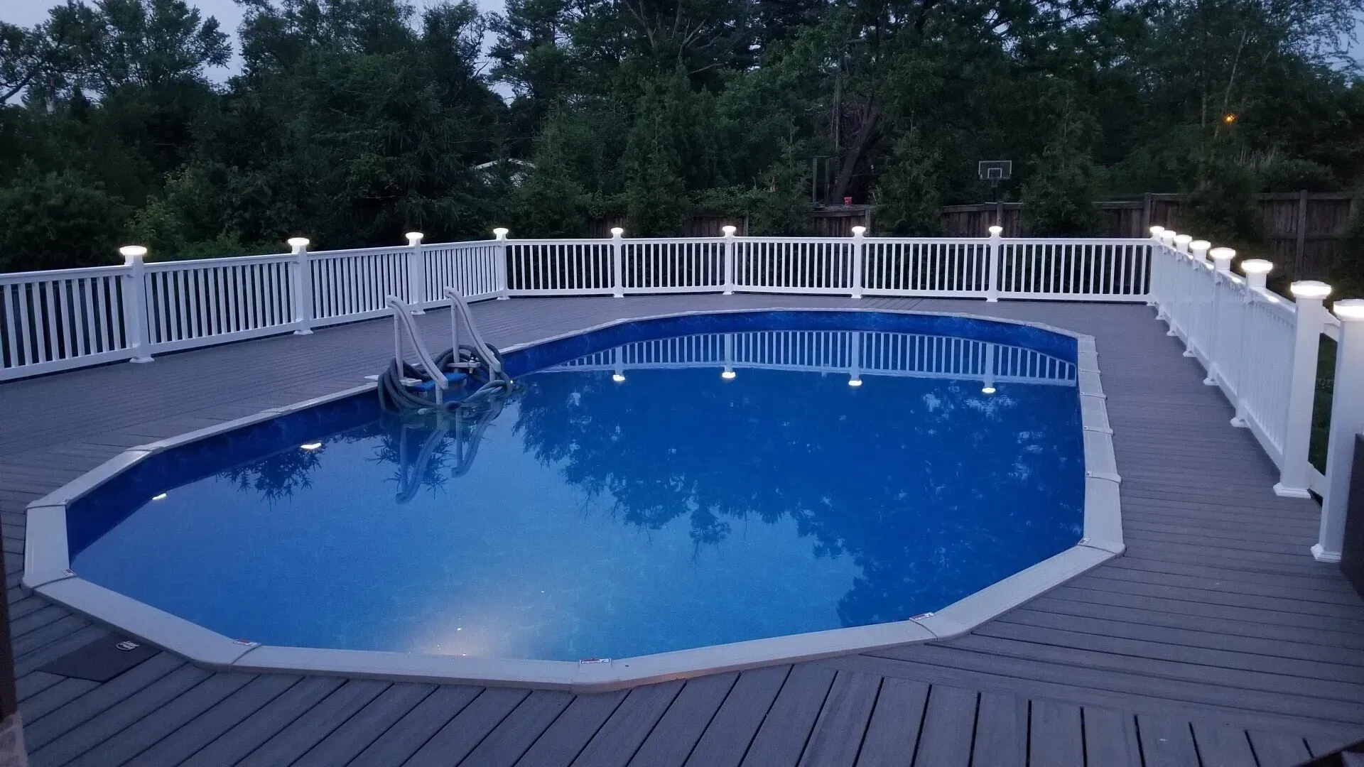Swimming pool surrounded by a gray deck and white fence with lights, set at dusk.
