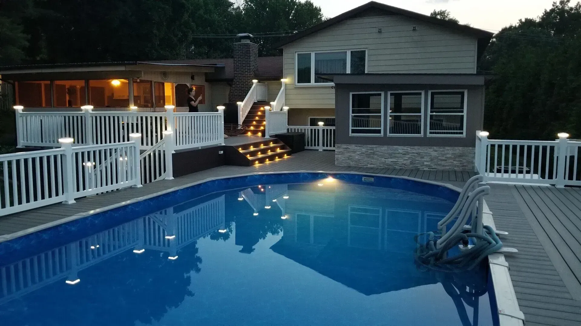 Backyard pool with house, deck, and stairs illuminated at dusk.