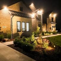 Night view of a house with bright exterior lights illuminating the brick facade and landscaping.