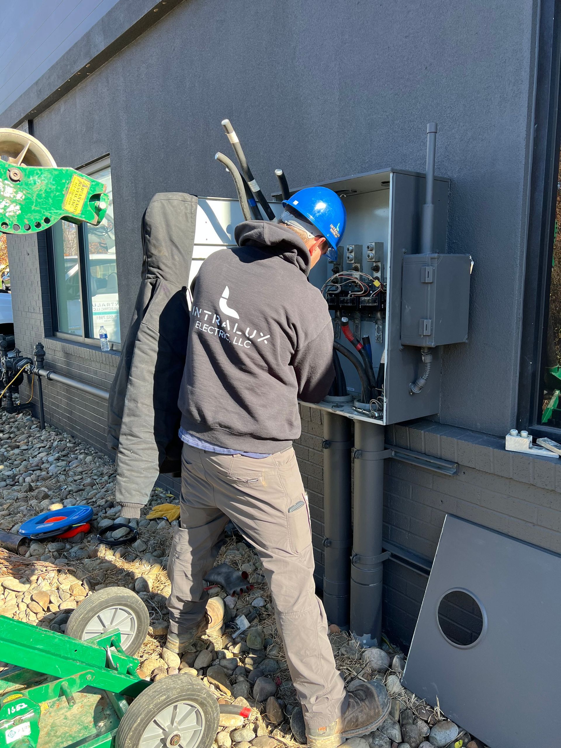 Electrician in blue hard hat working on an electrical panel outside a building with gray stucco.