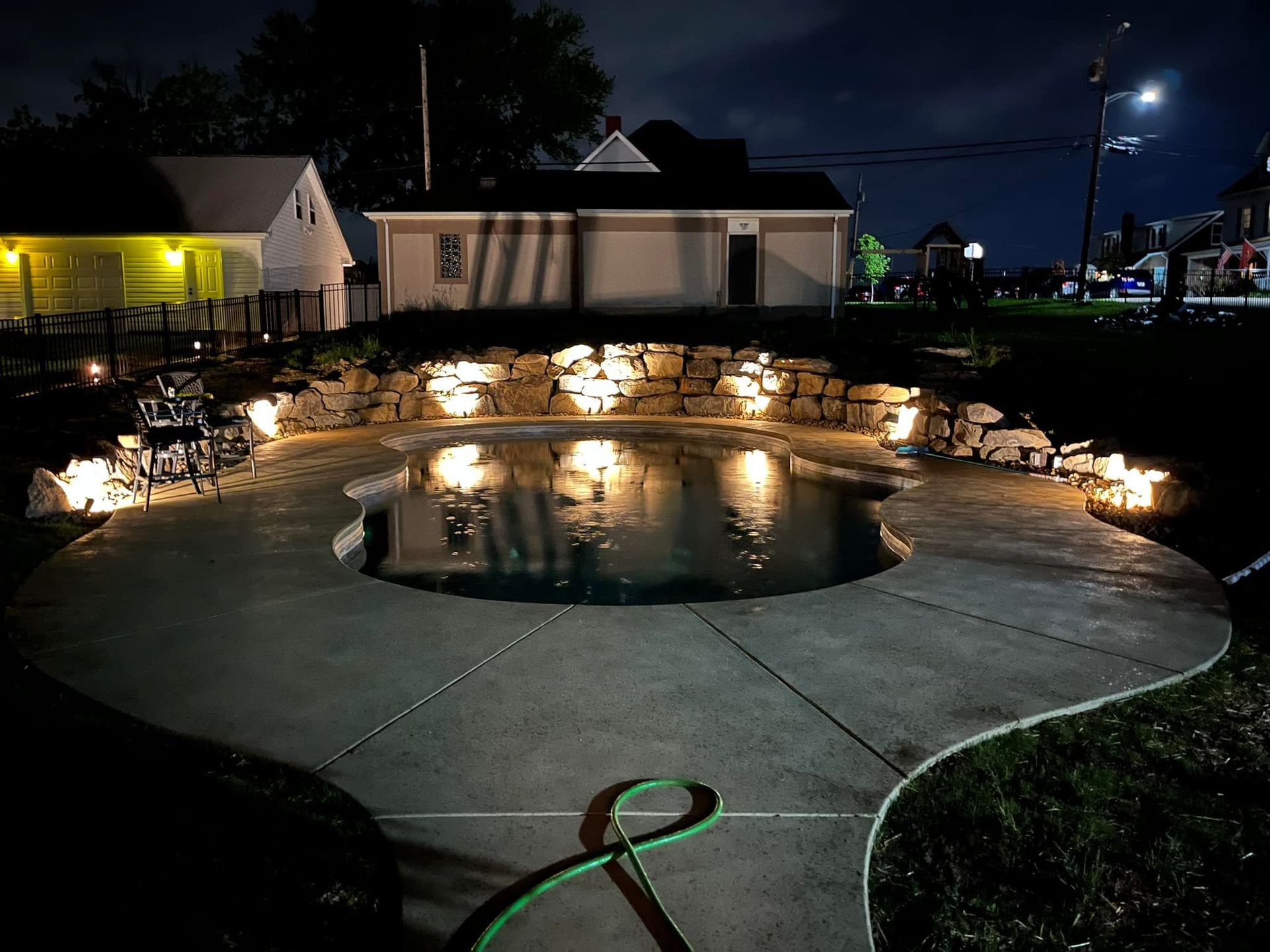 Nighttime view of a pool with surrounding stone wall and accent lights, in a yard with houses in the background.
