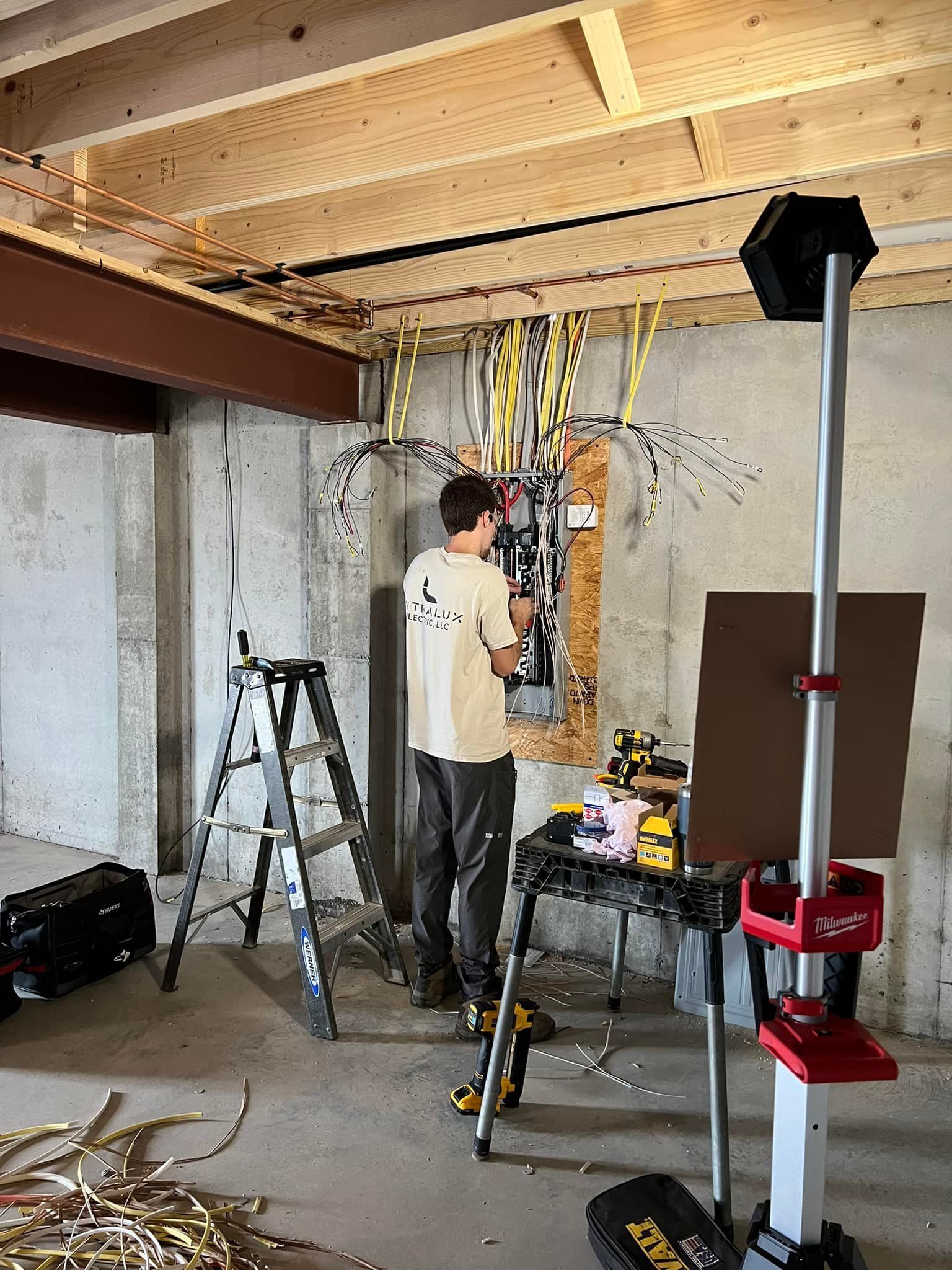 Man working on electrical panel in a basement. Wires, tools, and a ladder are present.