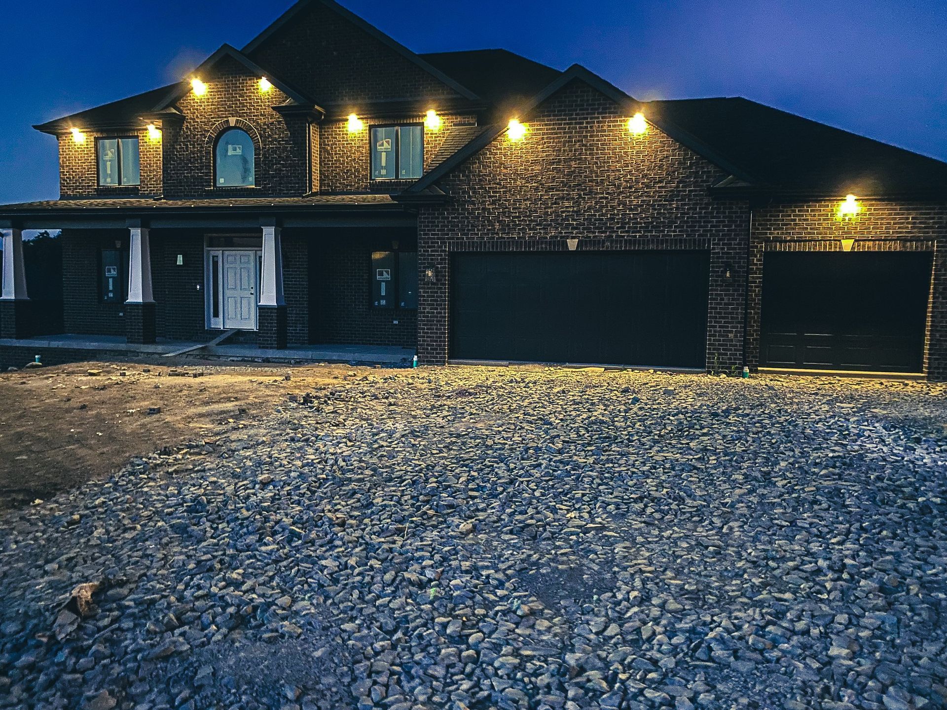Night view of a two-story brick house with lit garage and exterior lights, gravel driveway.
