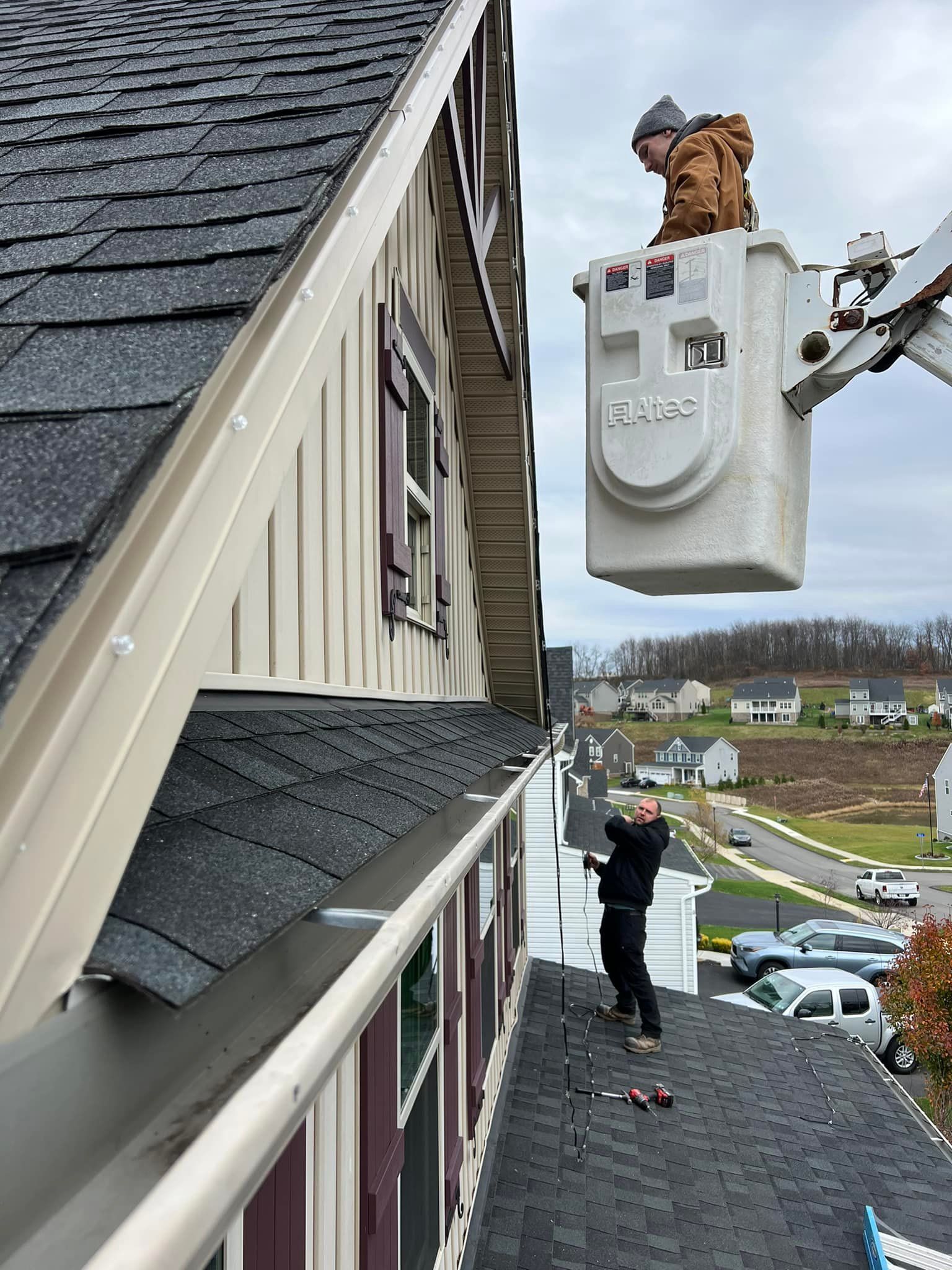 Two workers installing gutters on a house, one in a lift, one on the roof, overcast day.