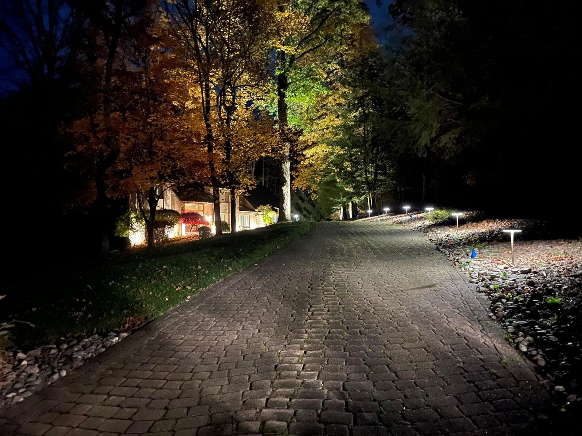 A brick driveway leads to a house with illuminated landscaping at night, flanked by trees with autumn foliage.