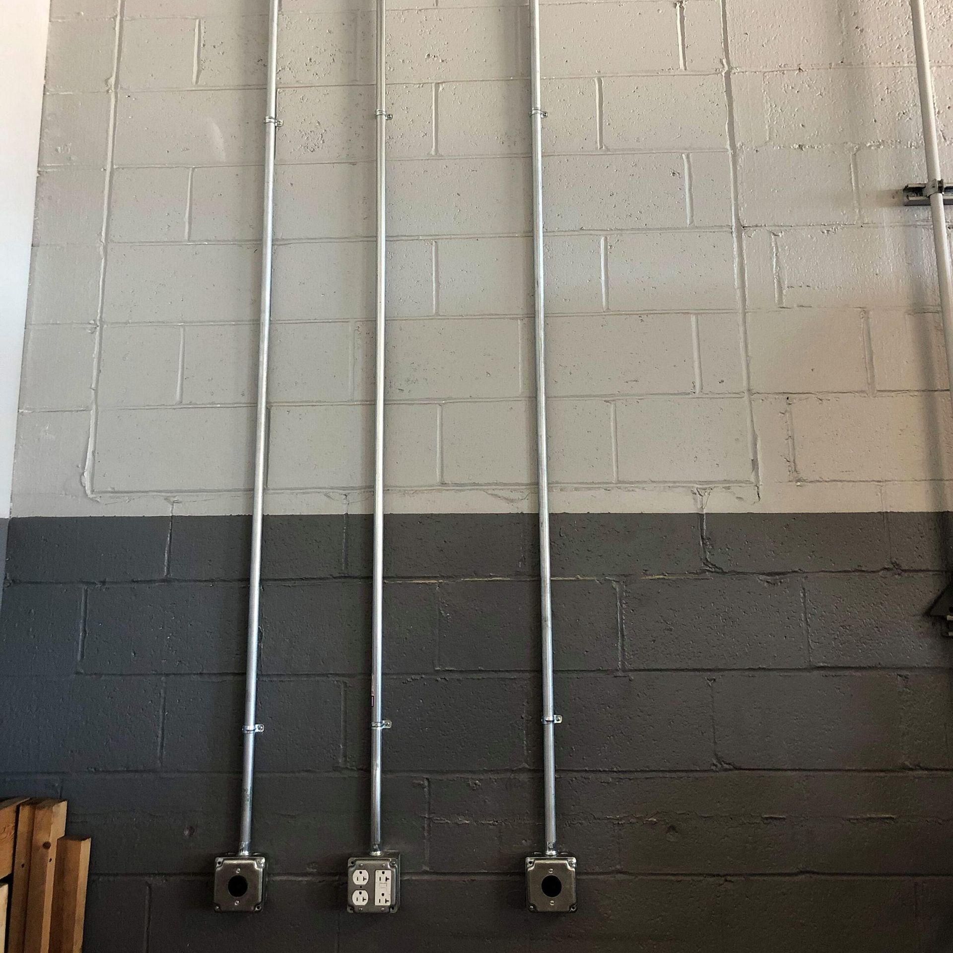 Three vertical metal conduits attached to a painted cinder block wall. Gray and white paint. Electrical boxes at the bottom.