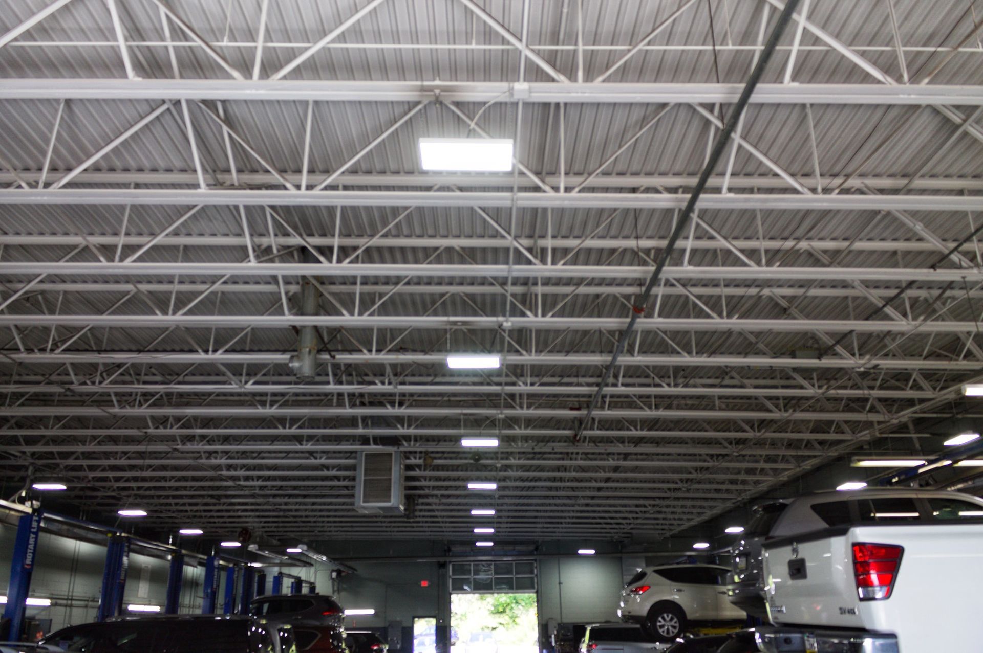 Interior of a vehicle repair shop with cars on lifts, looking up at a metal truss ceiling with lights.