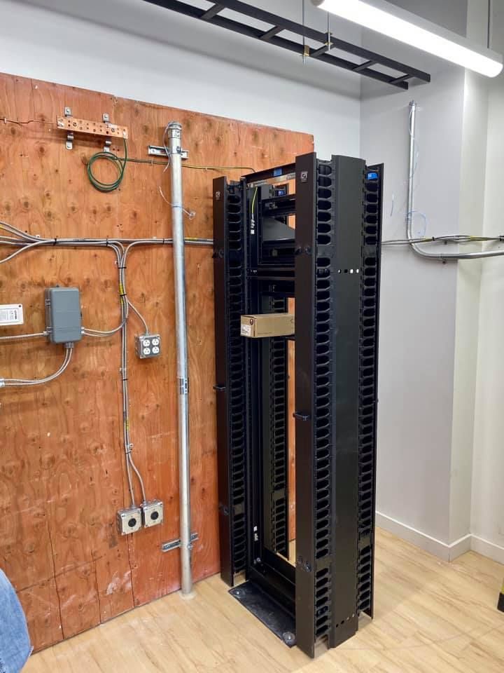 Black server rack next to a wooden wall with conduit and electrical boxes.