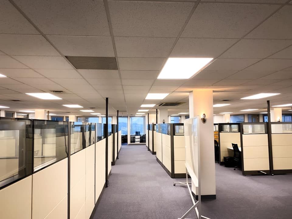 Rows of beige cubicles in an office setting. Fluorescent lights and a gray carpeted floor are visible.