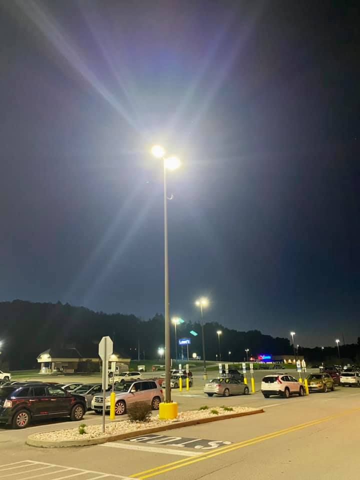 Parking lot at dusk with streetlights illuminating cars and a dark hillside in the background.