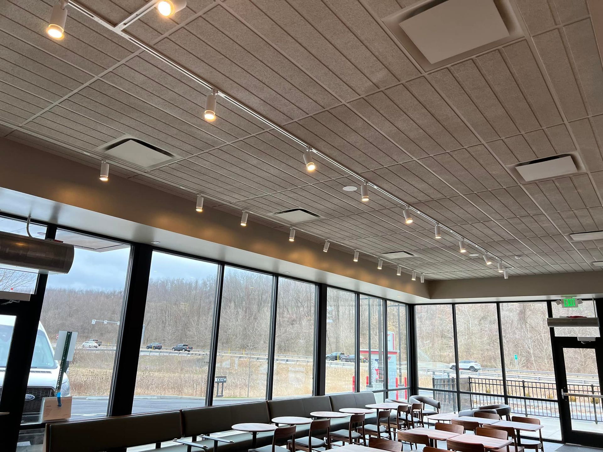 Interior of a brightly lit cafe with large windows overlooking a wooded area. Track lighting on the ceiling.