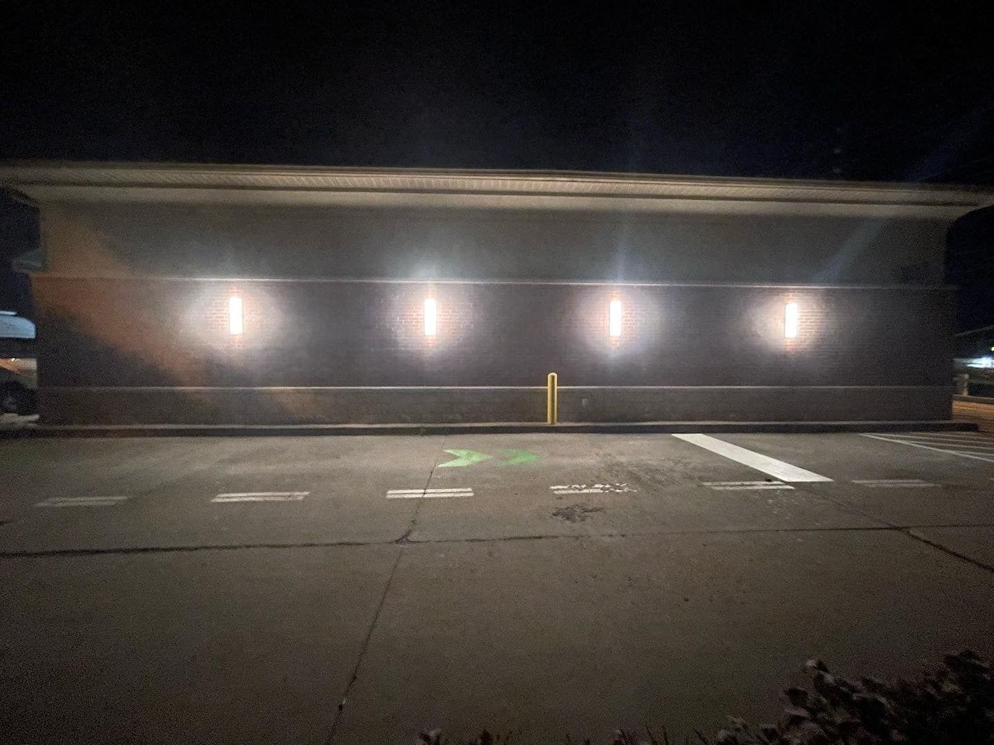 A long, dark concrete building exterior with four illuminated rectangles. A cracked asphalt parking lot in front.