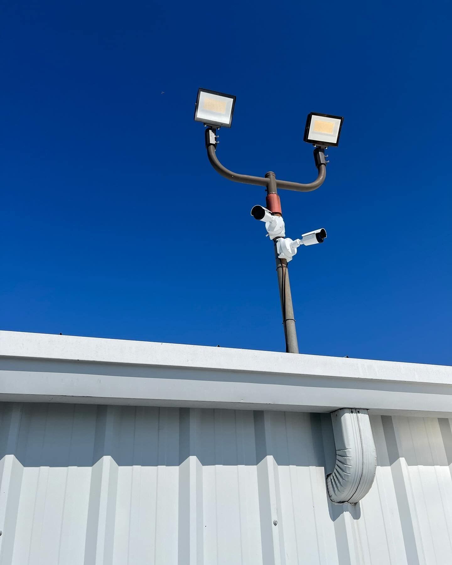 Light pole with two floodlights and security cameras against a blue sky.