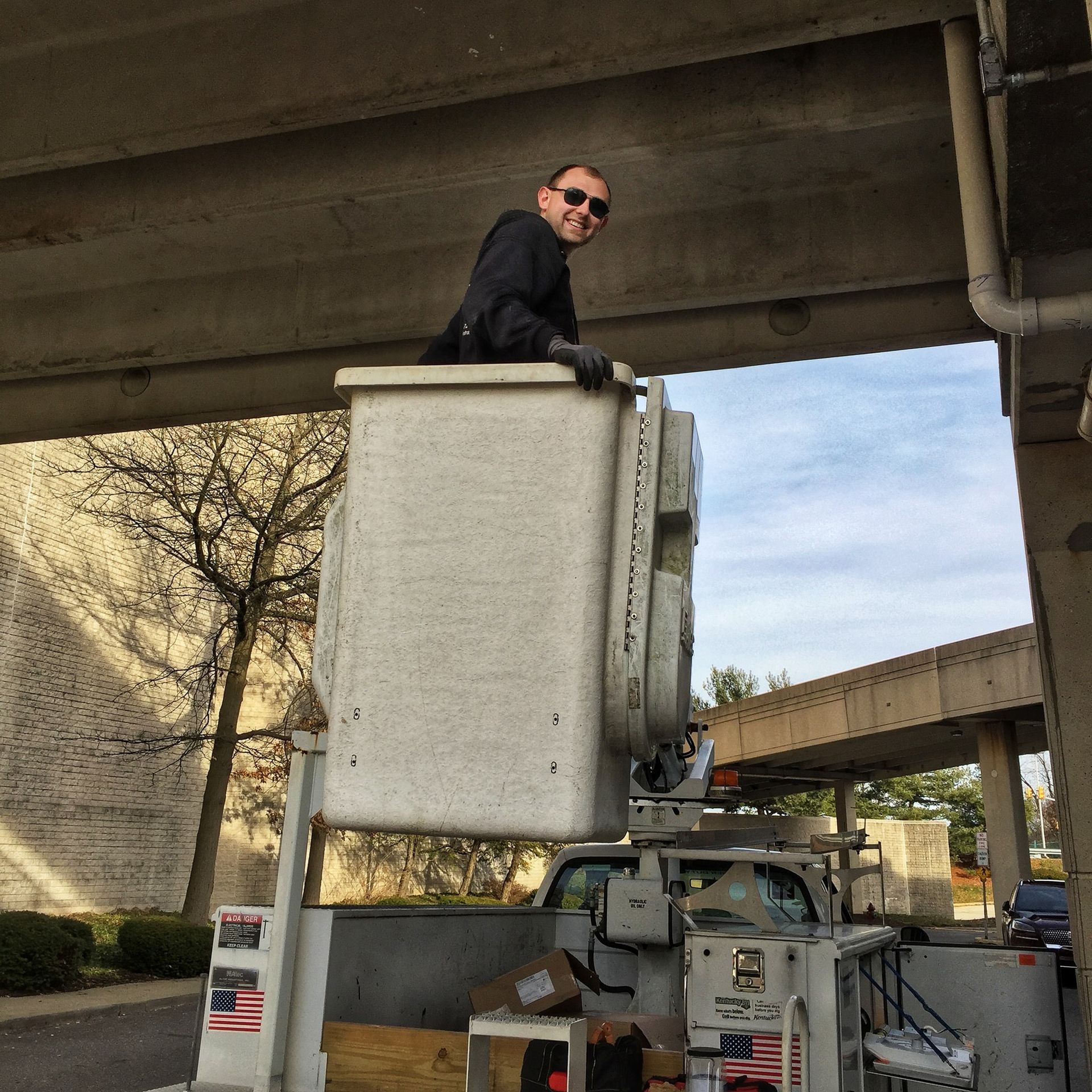 Man in bucket lift smiling, working on underside of concrete overpass. Day, outdoors.