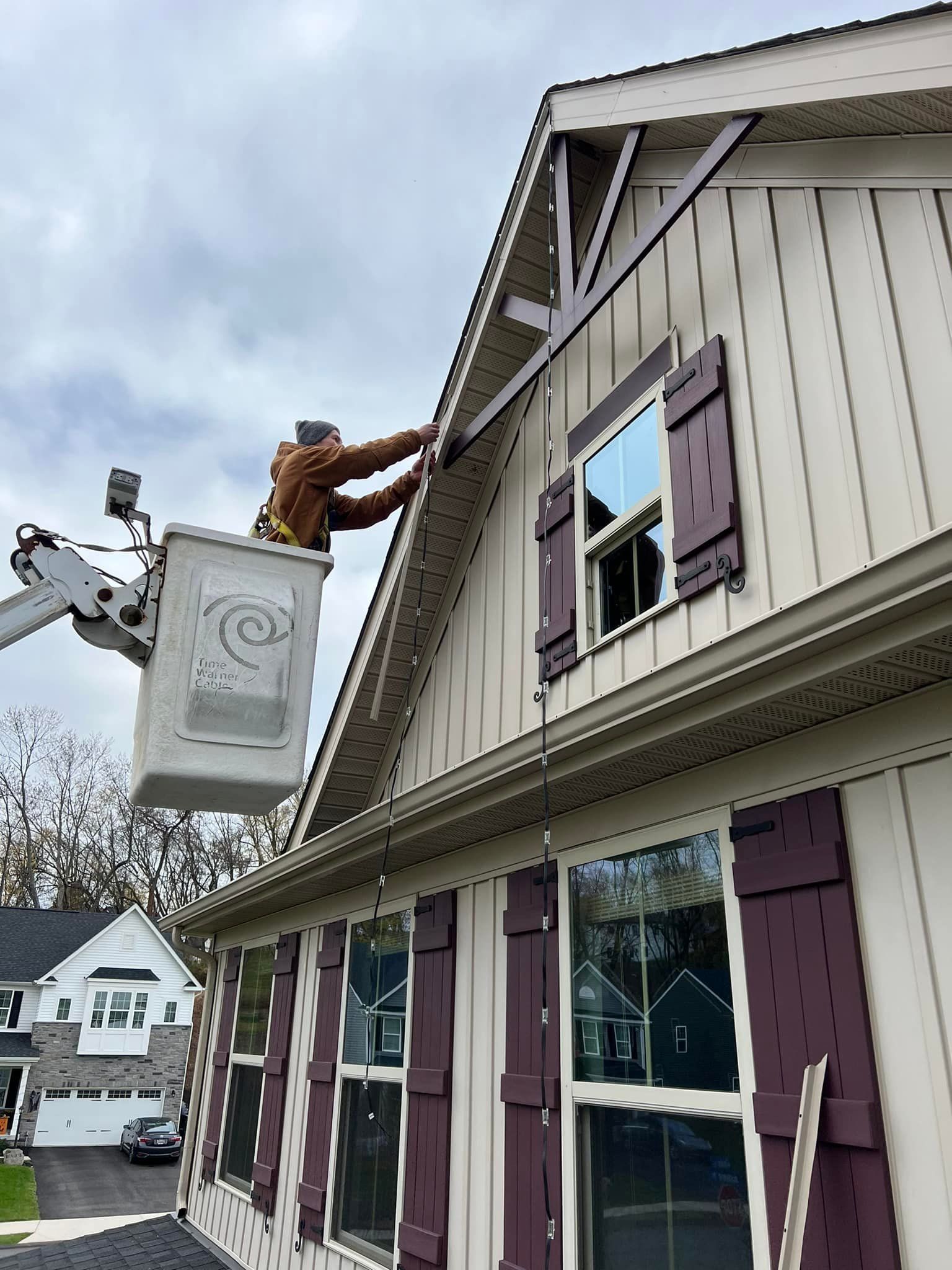 Person in a lift basket installing something on the roof of a house with beige siding and purple shutters.