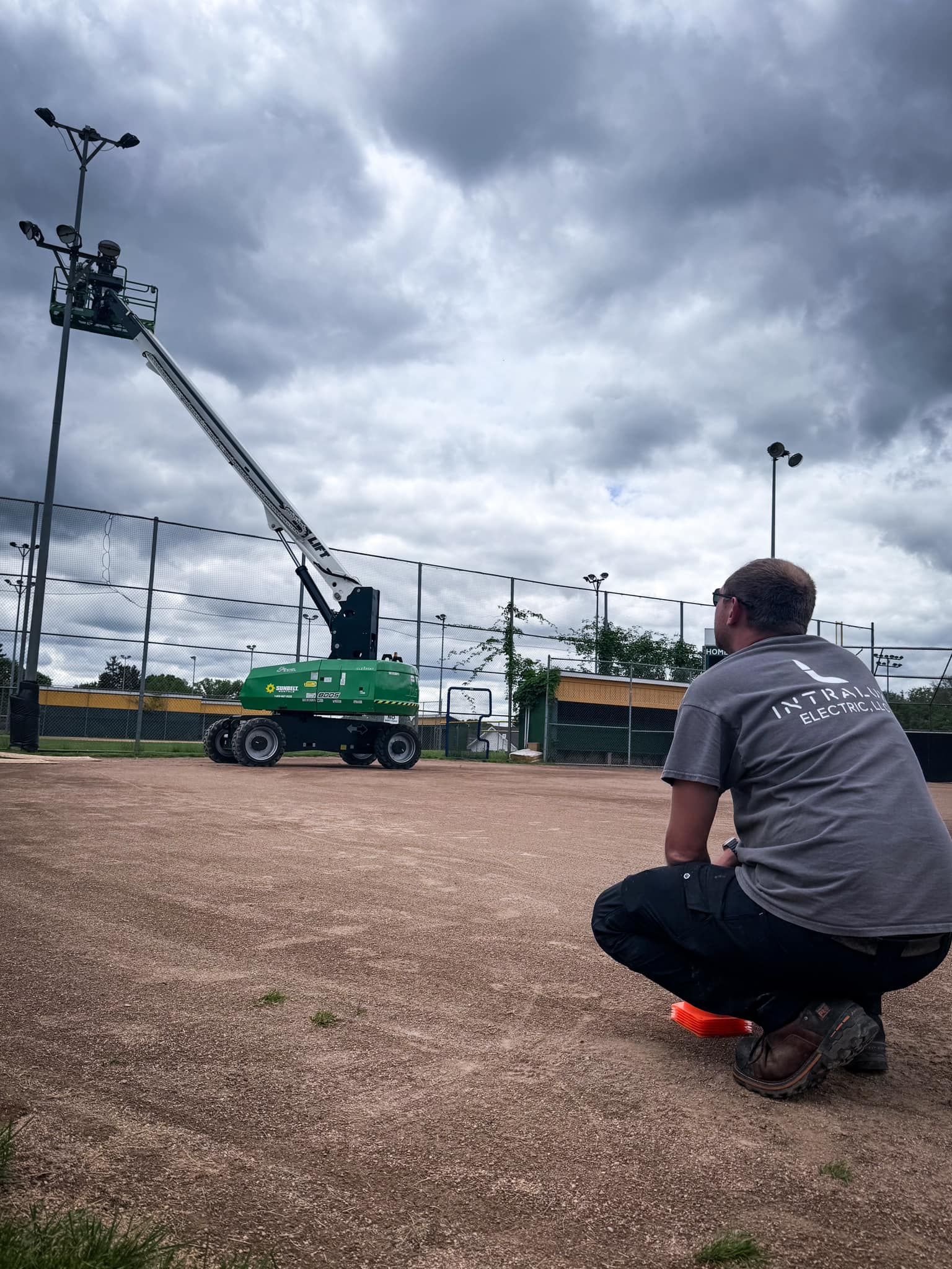 Person on a baseball field watches a lift working on a light pole under a cloudy sky.