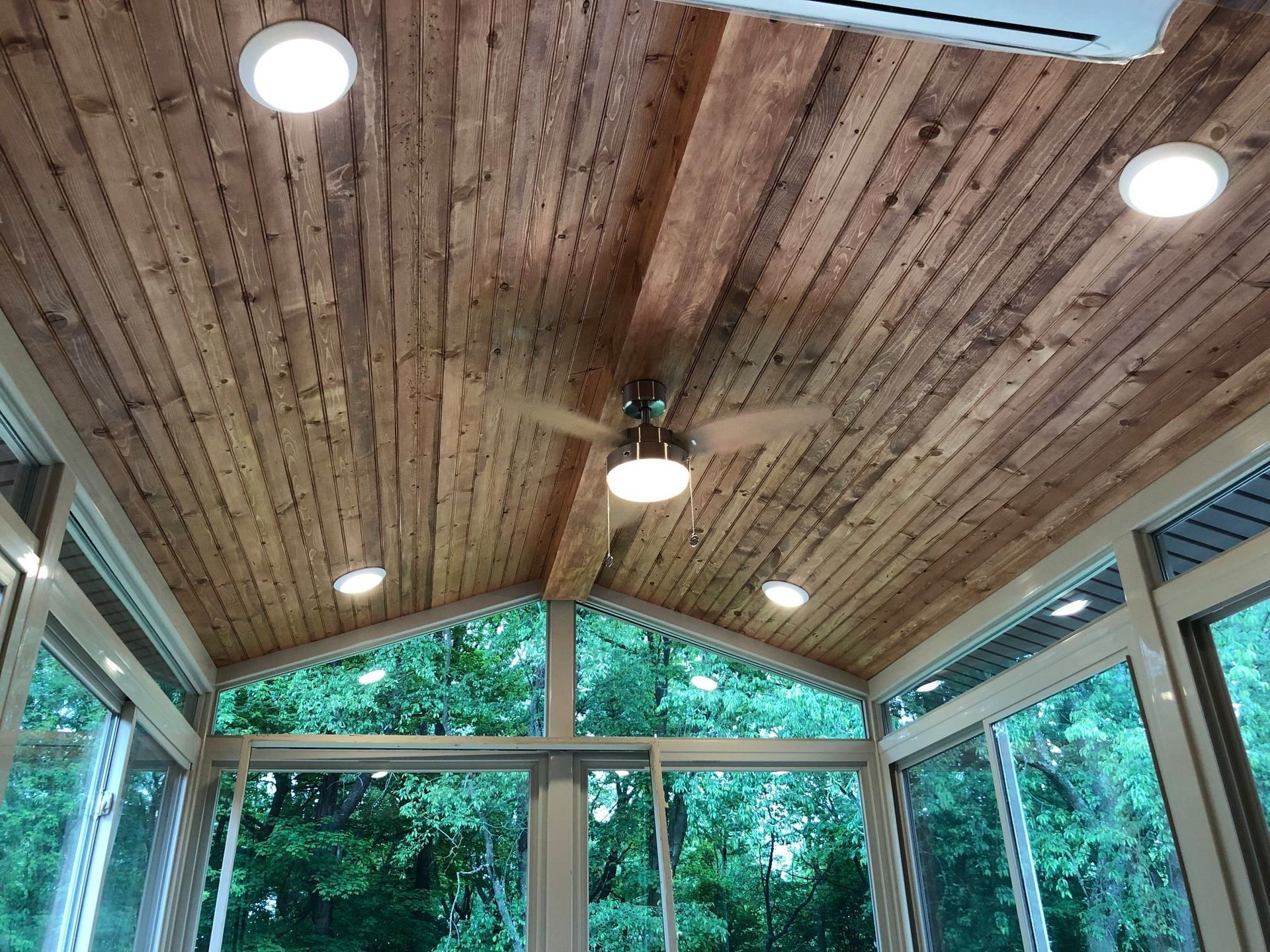 Sunroom with wooden ceiling, recessed lights, fan, and large windows looking out to greenery.