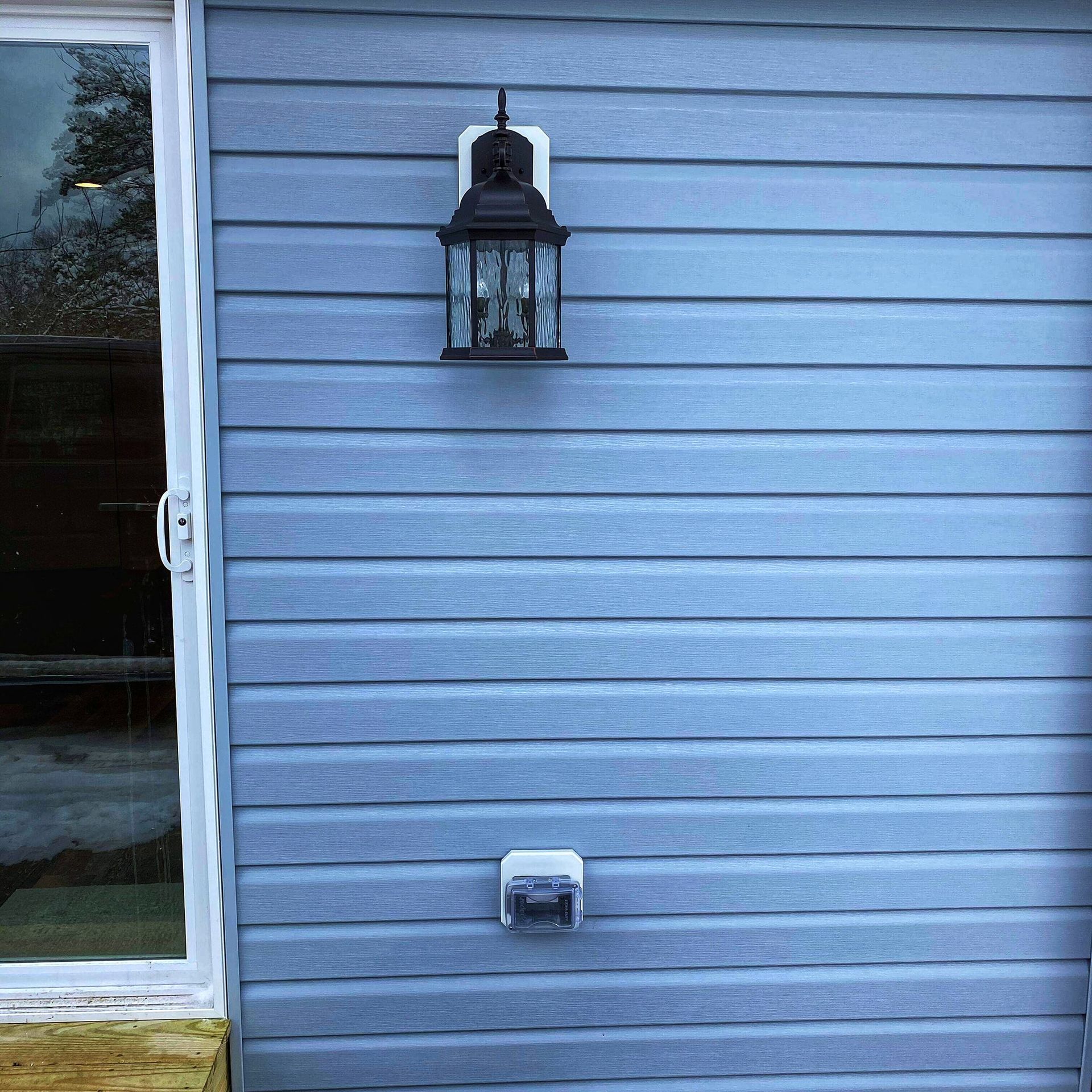 Blue siding of a house with an outdoor lantern and an electrical outlet next to a sliding glass door.
