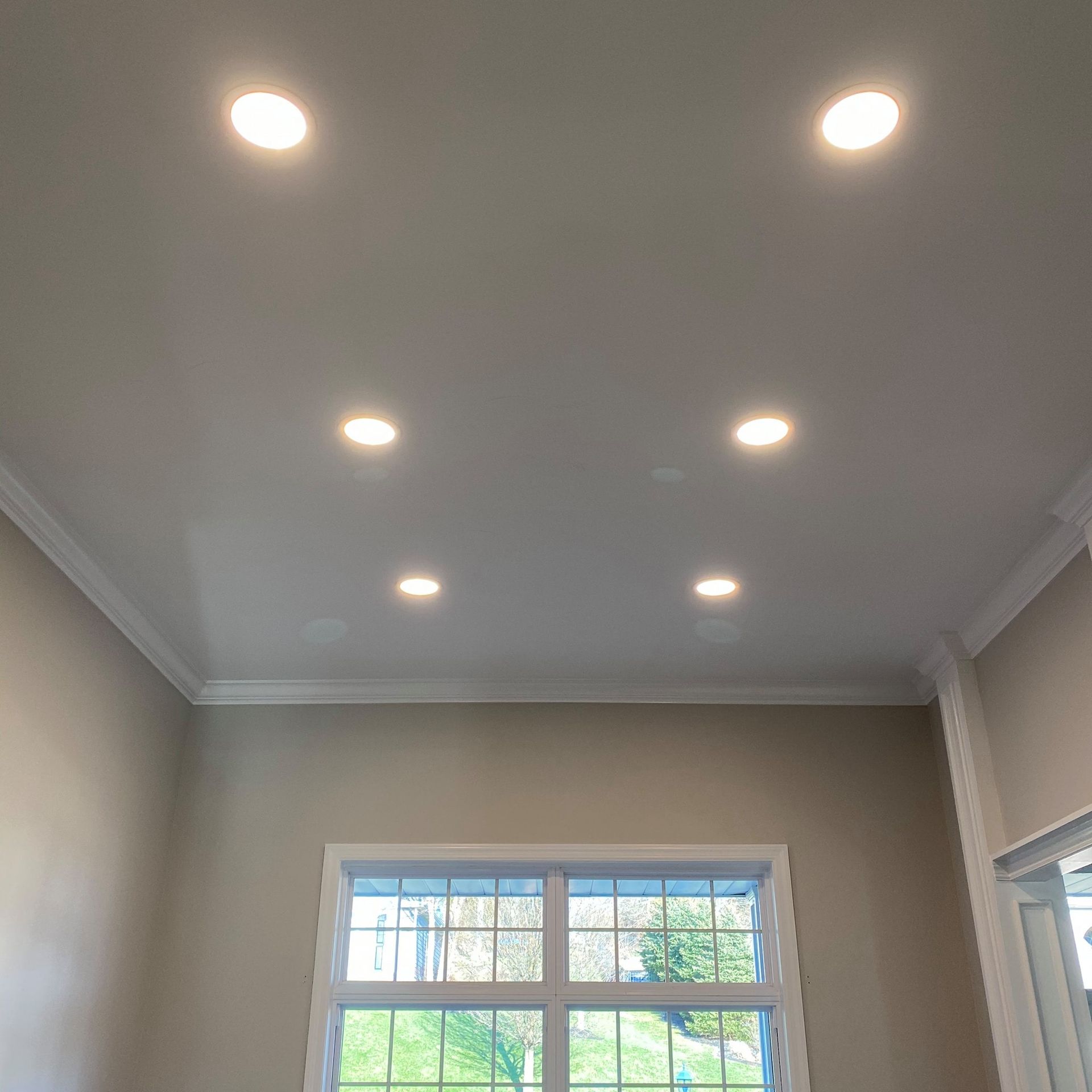 Ceiling with six recessed lights and crown molding over a window and tan walls.