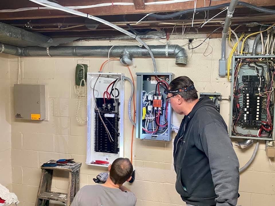 Two electricians work on electrical panels in a basement setting.