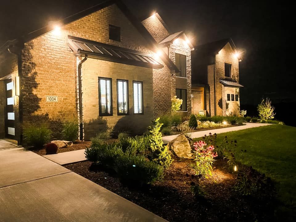 Nighttime view of a brick house illuminated by outdoor lighting; landscaped yard, paved walkway, dark sky.