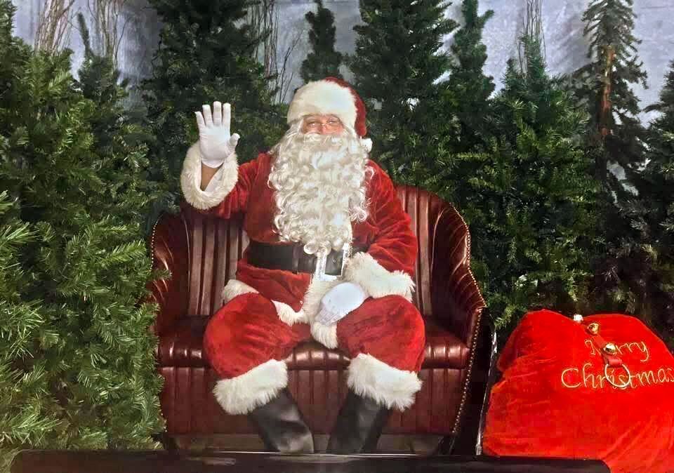 Santa claus is sitting on a couch waving at the camera.