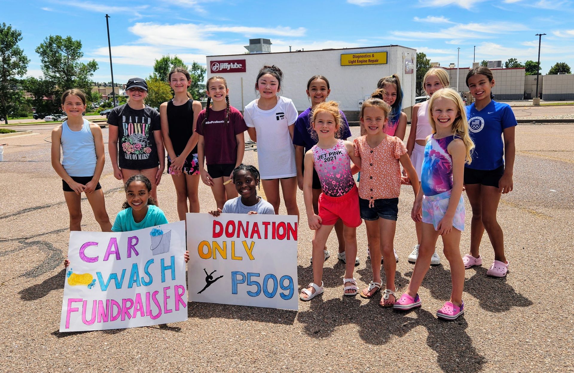 Group of young girls holding signs for a car wash fundraiser in a parking lot, with a food truck in the background.