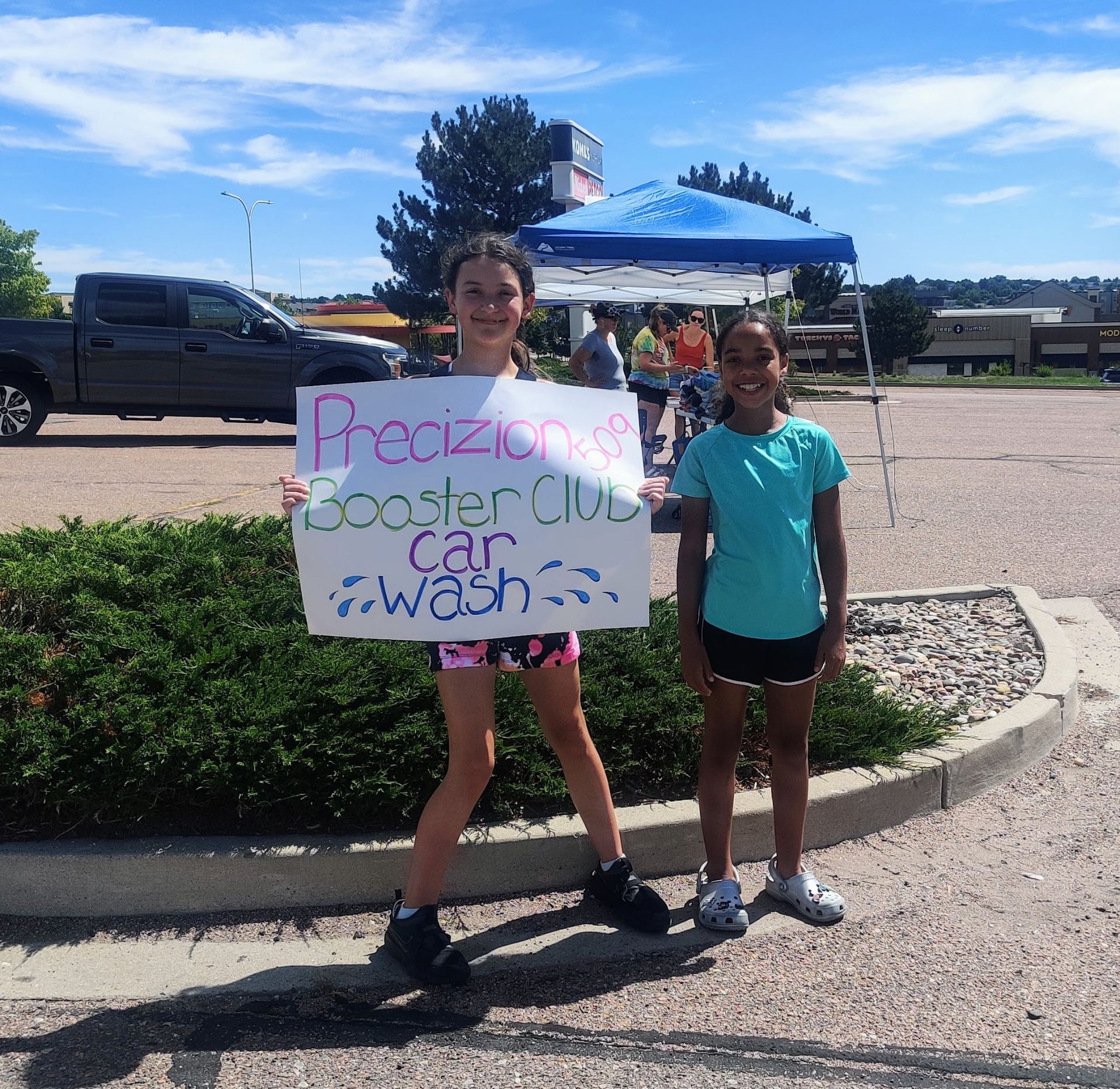 Two young girls holding a sign for a car wash, standing outside under a tent on a sunny day.