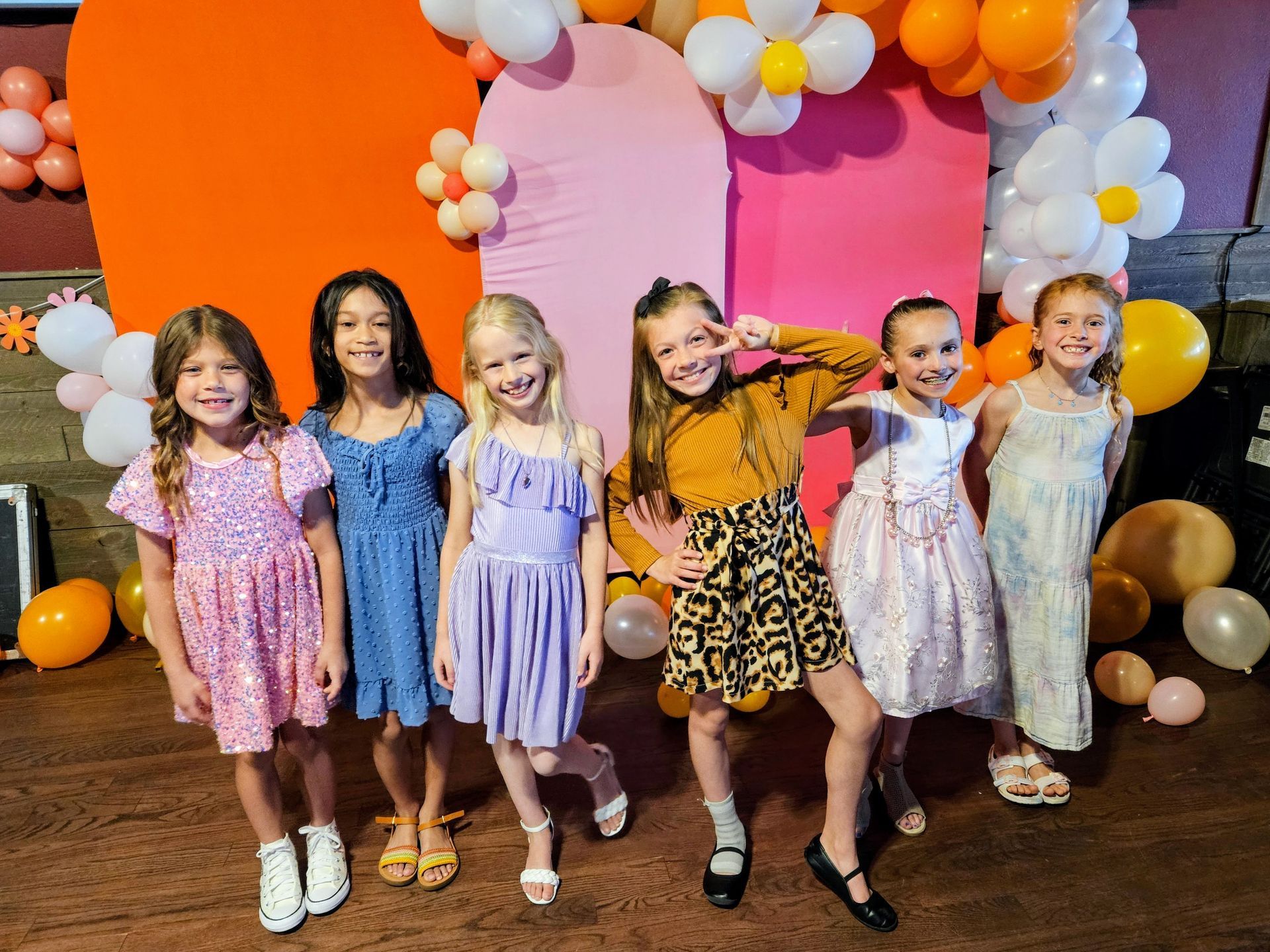 Six girls pose in front of a colorful backdrop with balloons. The girls wear dresses and have smiling expressions.