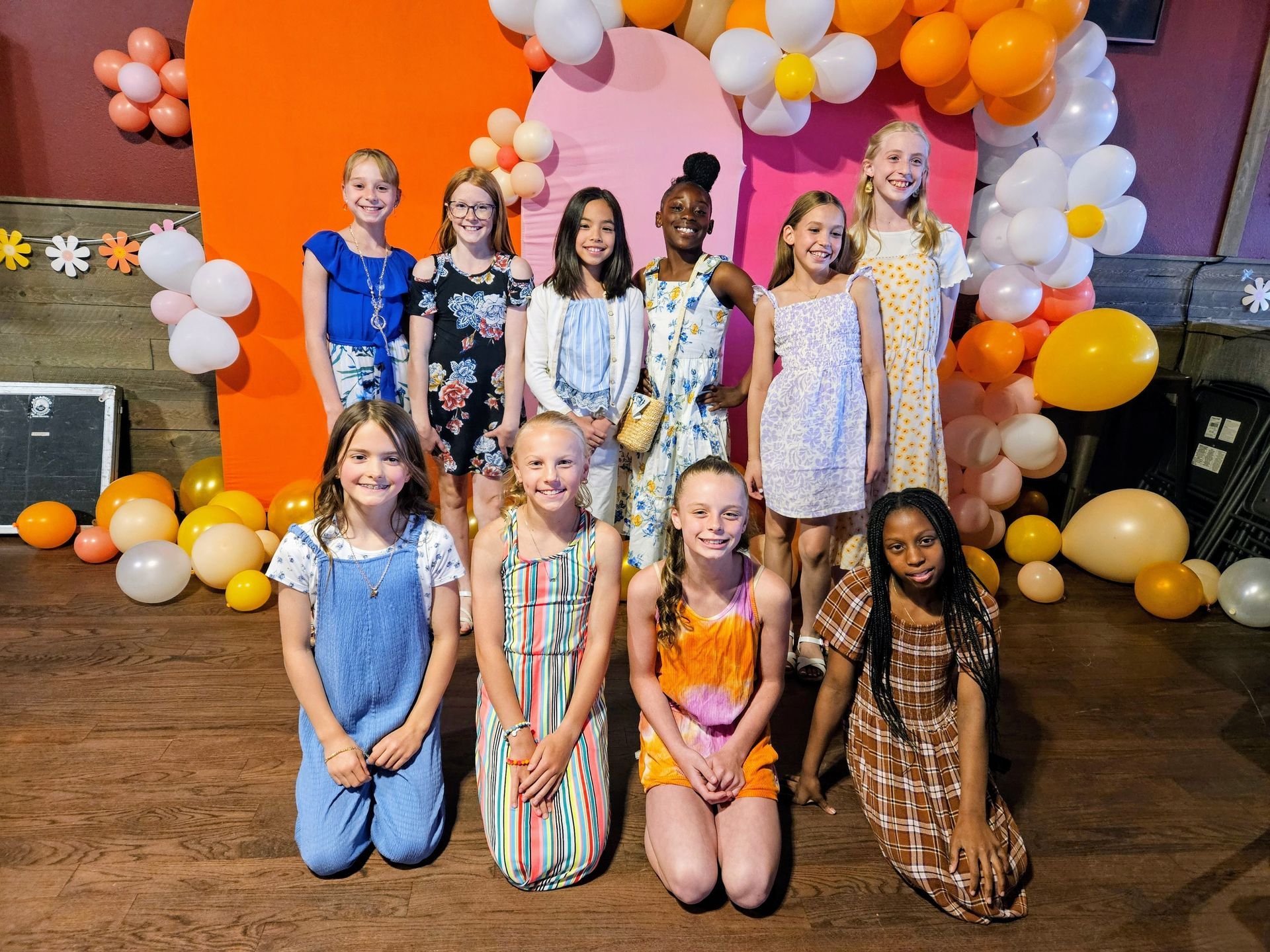 Group of eleven young girls posing in front of an orange and pink decorated backdrop with balloons. They're smiling and wearing colorful outfits.