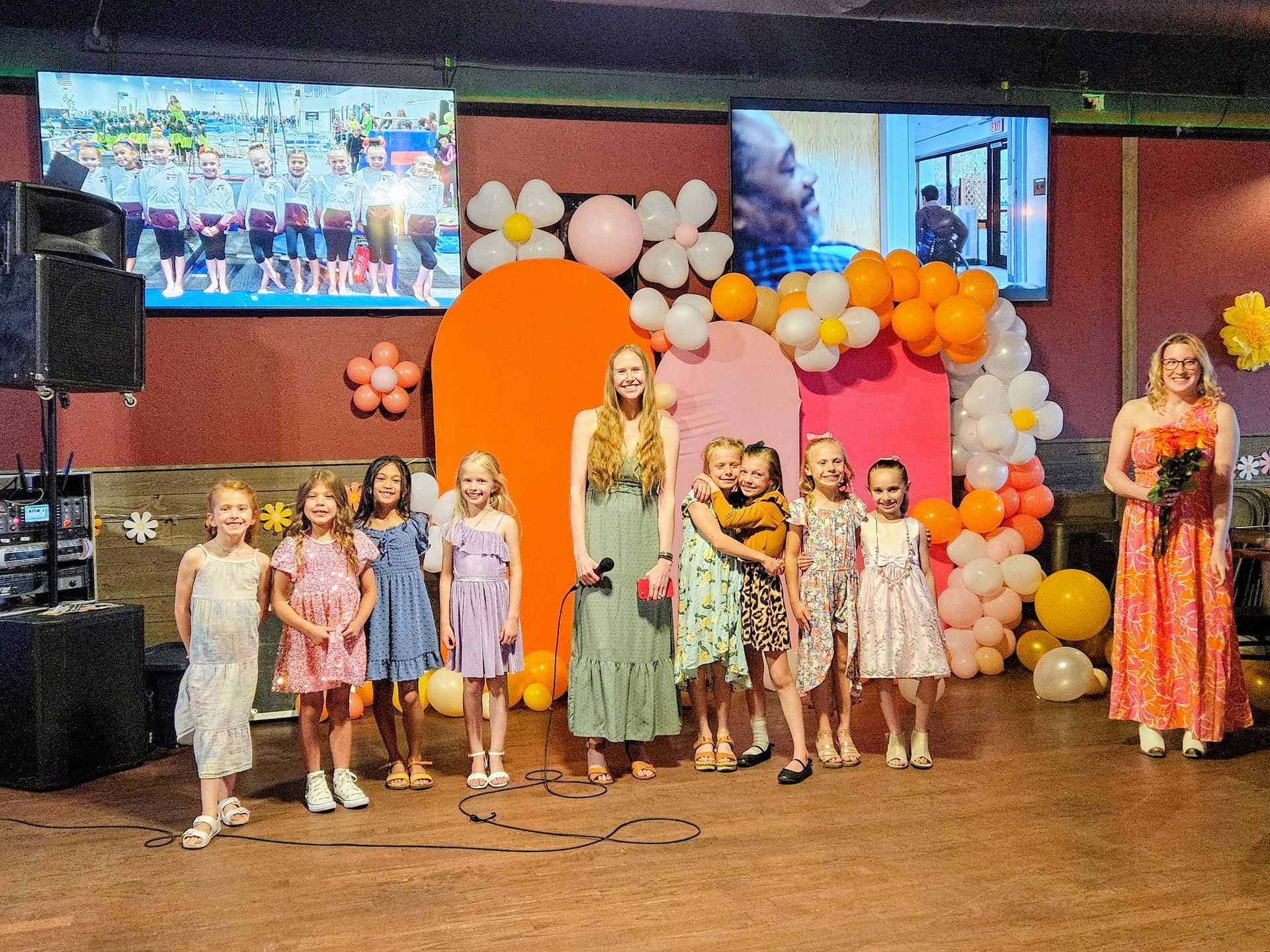 Group of children and women on a stage decorated with balloons and flowers. Two women hold microphones. Two screens display video.