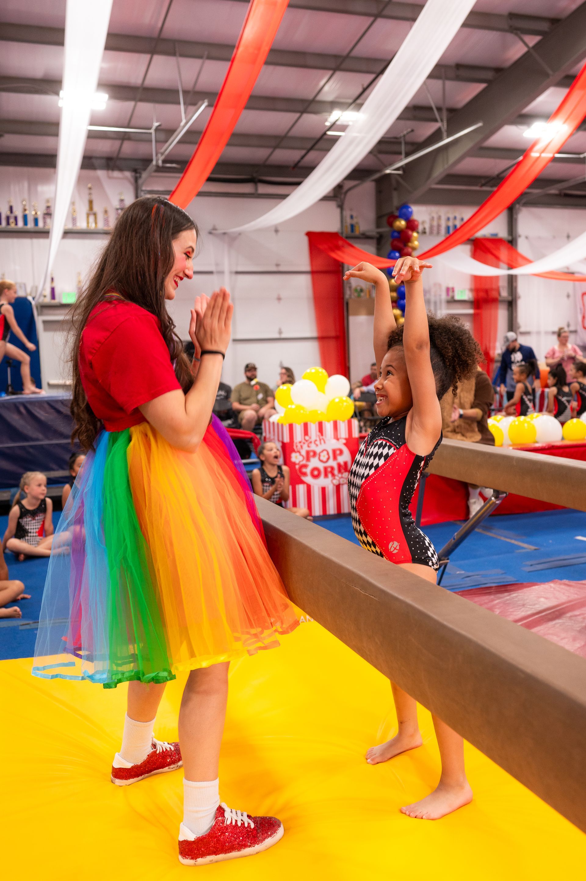 A girl in gymnastics attire raises her arms on a balance beam while smiling at a woman clapping. They are inside a gym decorated with red and white.