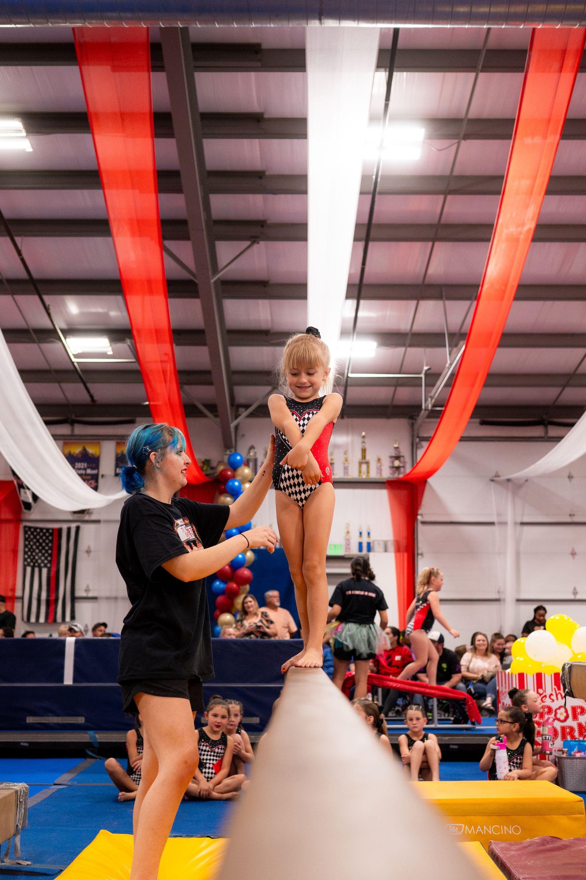 A young girl balances on a beam, assisted by a coach with blue hair, in a gym decorated for an event.