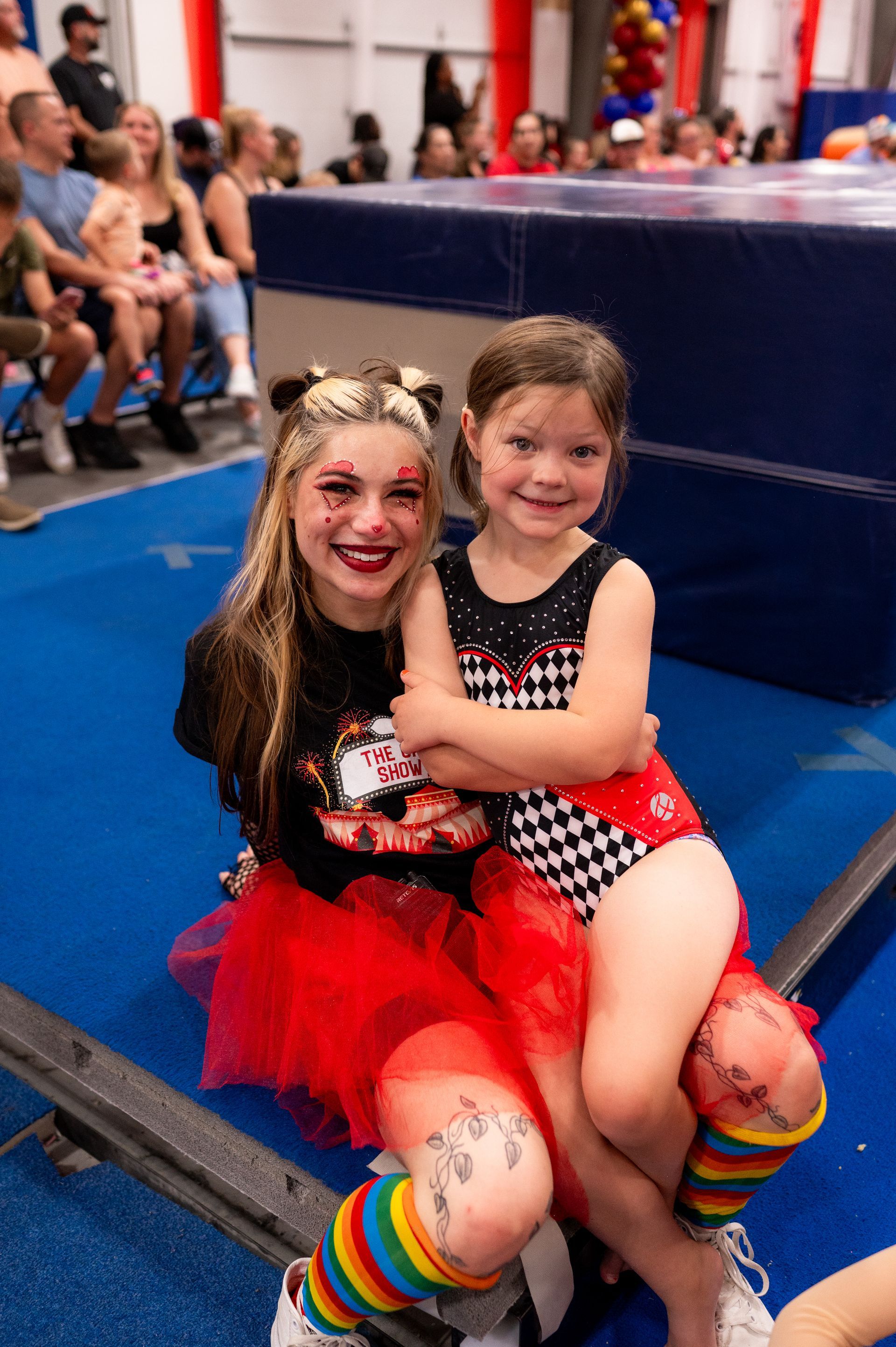 Two girls, one in a tutu, embrace on a blue mat. One has rainbow leg warmers and face paint. They are smiling in a gymnasium.