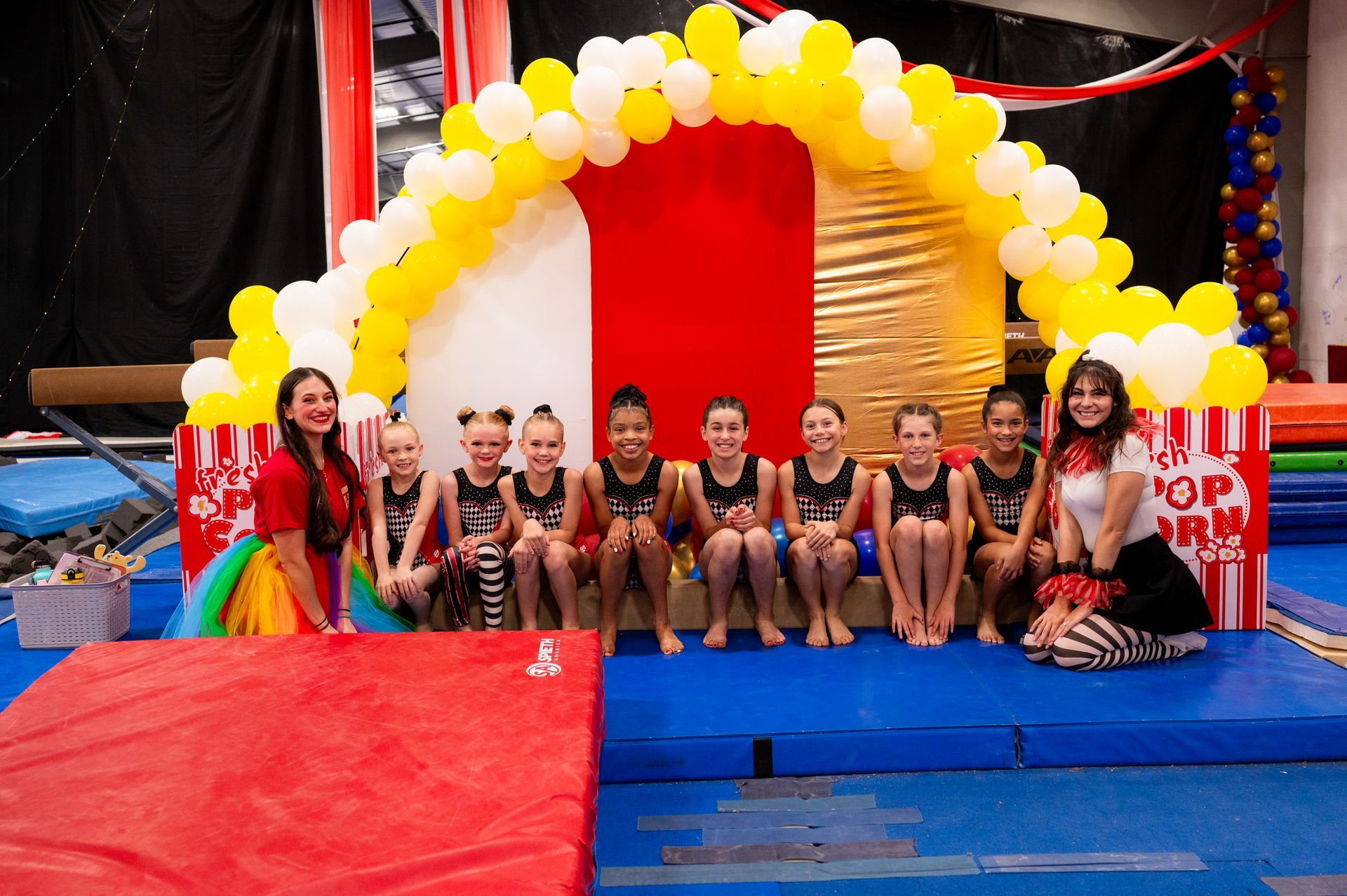 Group of gymnasts and coaches posing in front of a circus-themed backdrop with popcorn decorations. They are smiling in a gym setting.