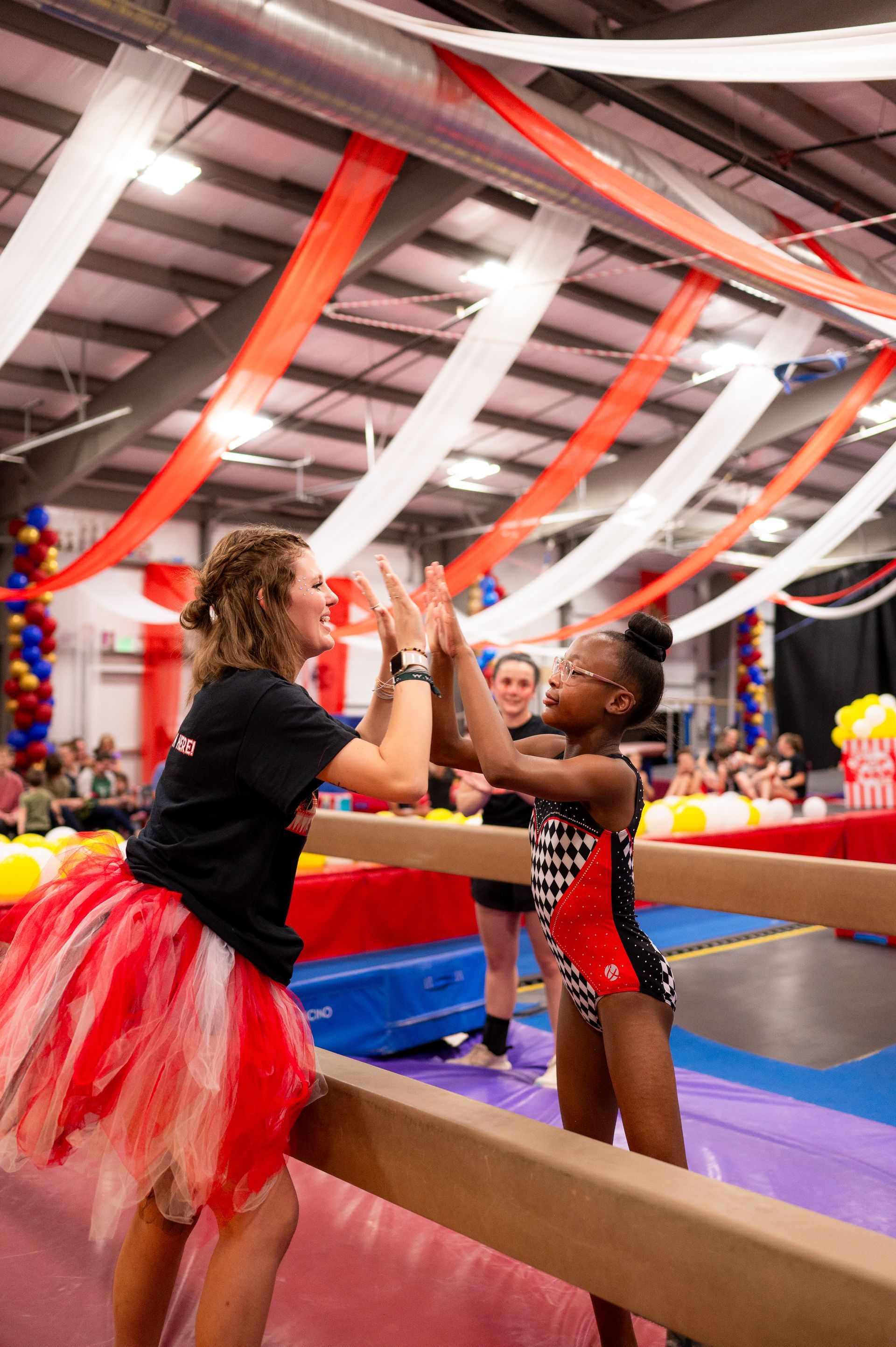 A gymnast high-fives her coach after completing a routine on a balance beam. The gym is decorated with red, white, and blue streamers.