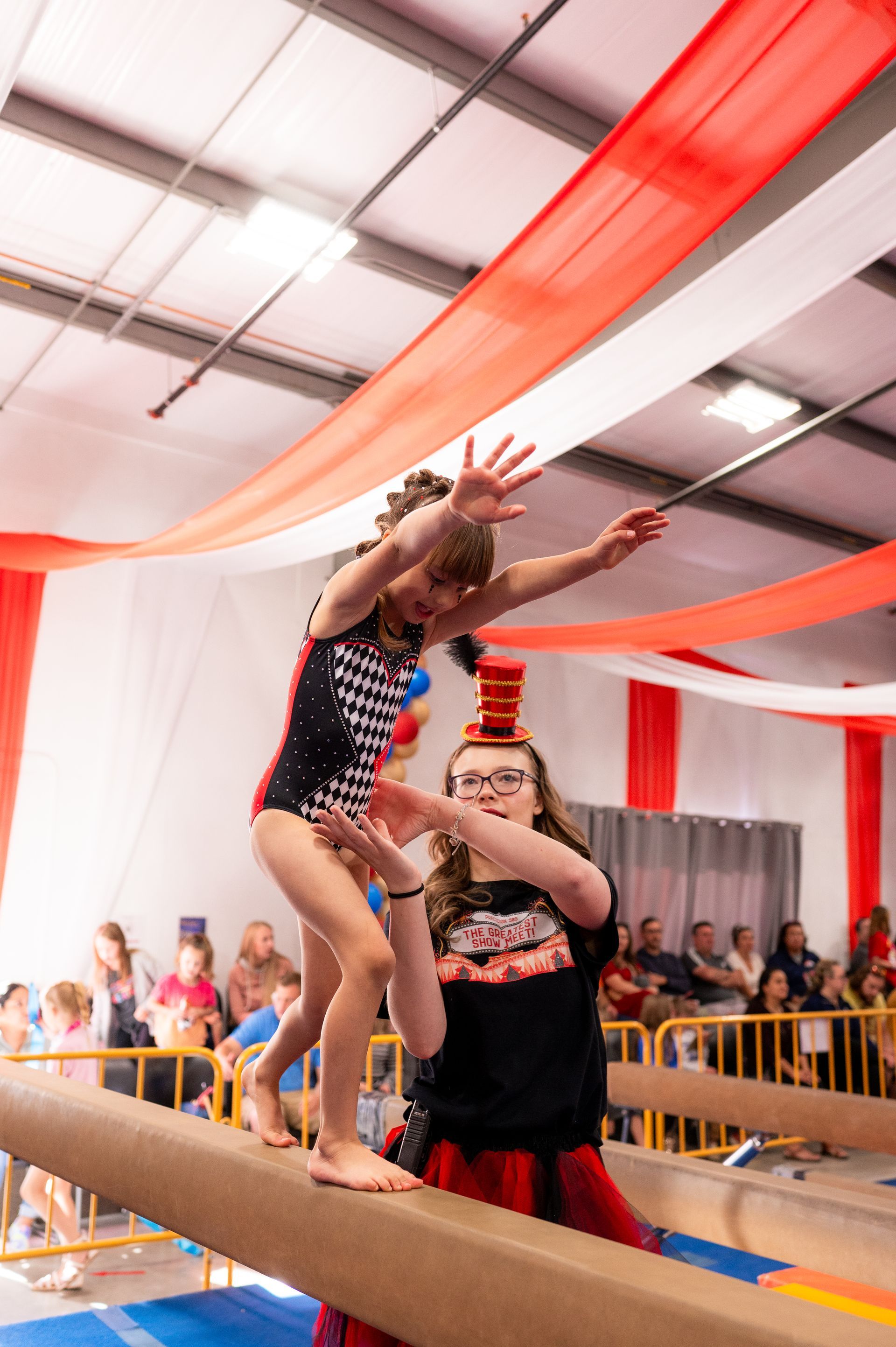A young gymnast balances on a beam, assisted by a coach in a gym decorated with red and white streamers.