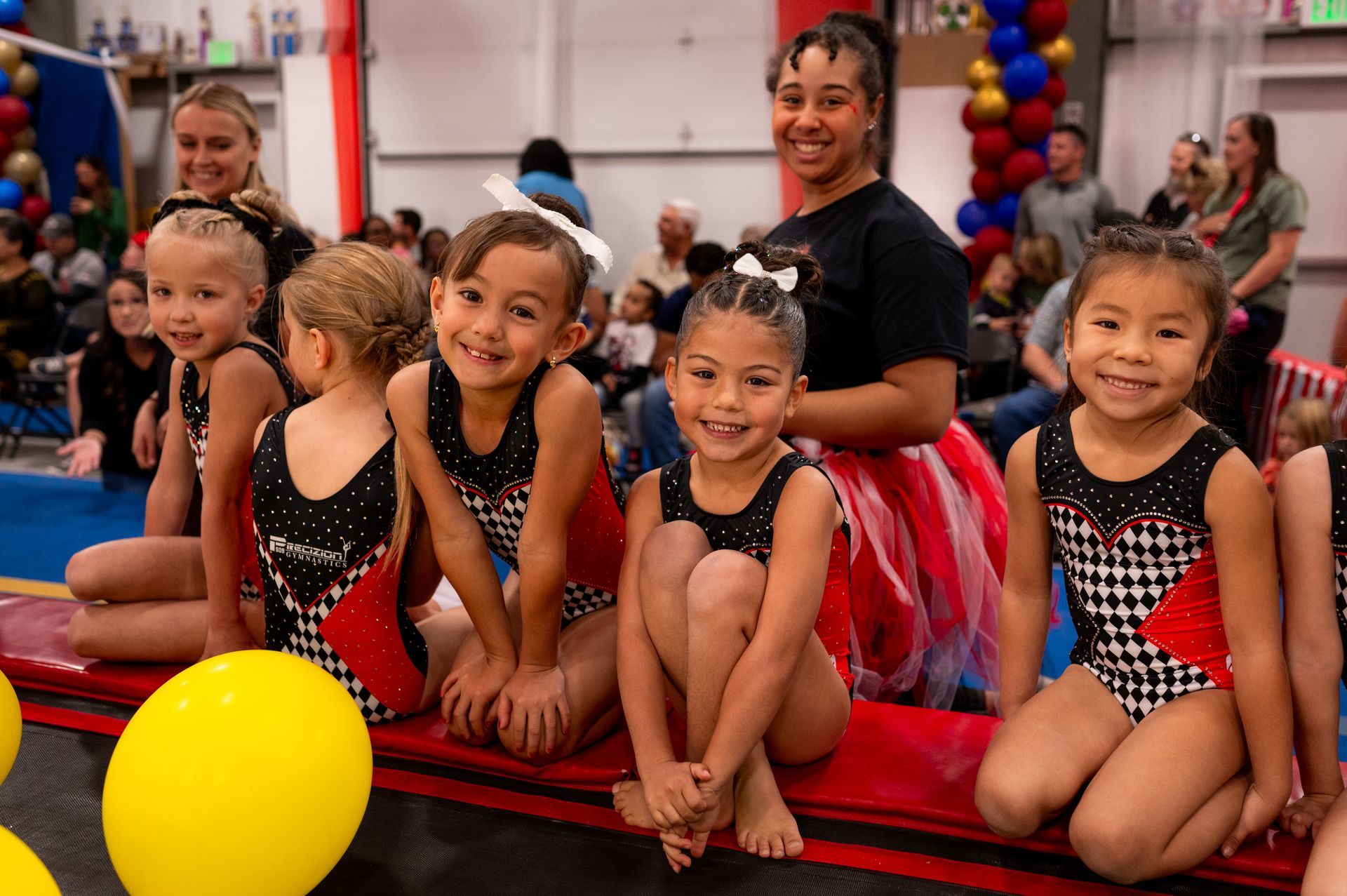 Young gymnasts in leotards smile for the camera, sitting with coaches on a red platform. Balloons and spectators fill a gymnasium.