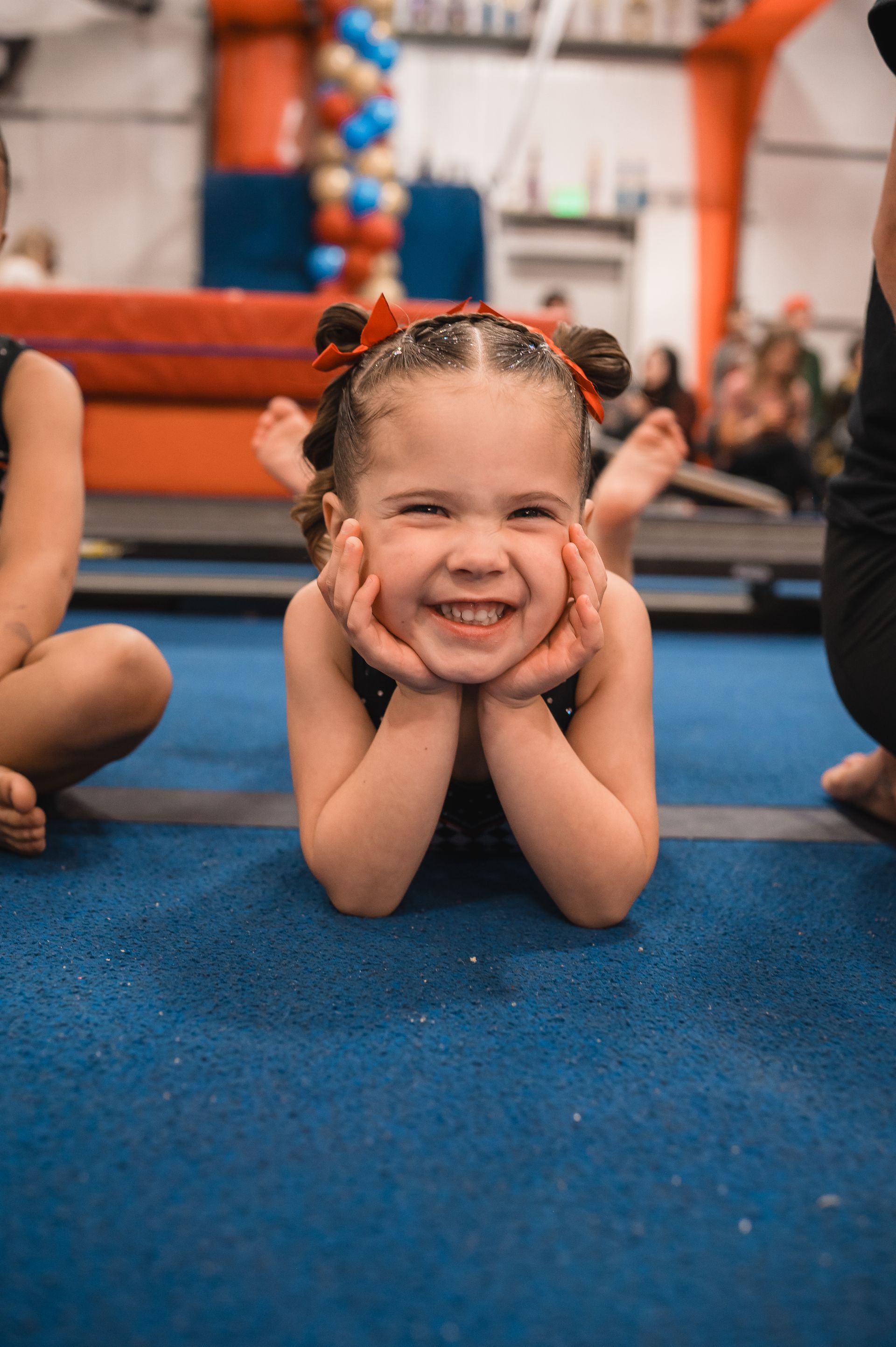 Young girl with pigtails and red bows smiles while lying on blue gym floor, hands under her chin.