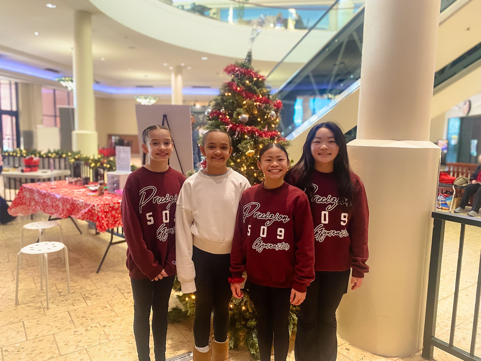 Four young girls in burgundy sweatshirts pose in front of a Christmas tree in a mall setting.