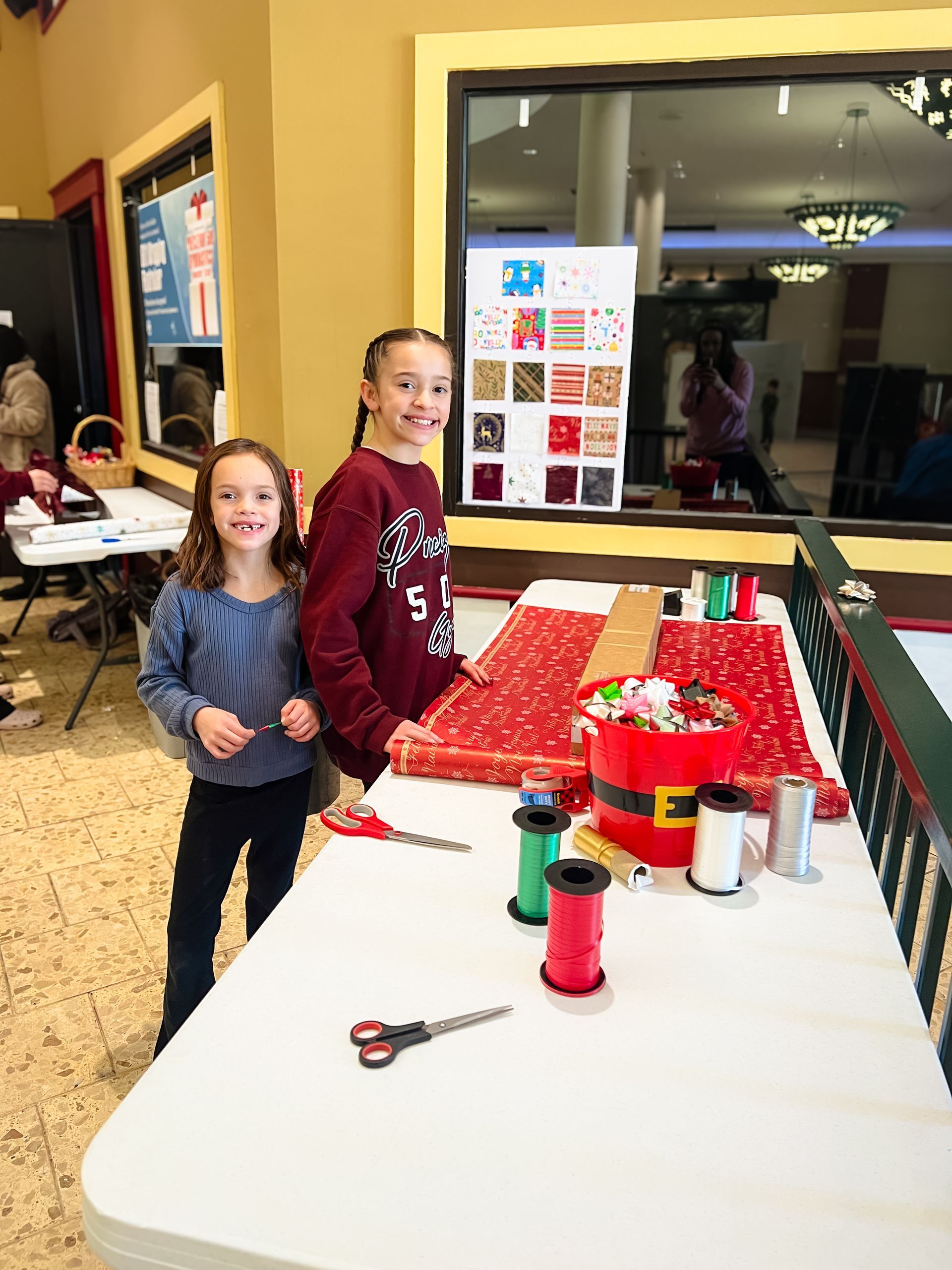Two girls at a gift-wrapping station, smiling. One wears a blue shirt, the other a maroon sweater. There's wrapping paper, scissors, and ribbon on the table.